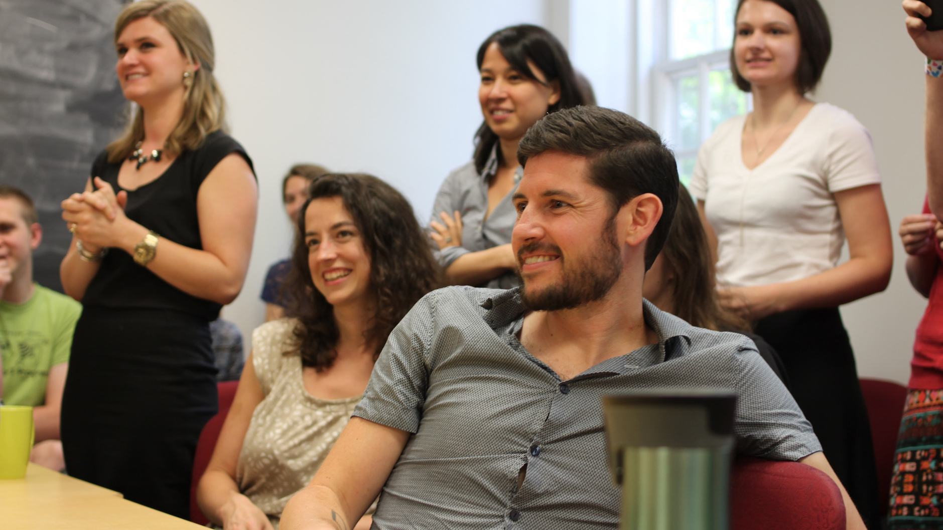 Aaron White, a PhD student in Linguistics, surrounded by faculty and classmates, smiling as he watches a colleague be congratulated on a successful defense of her dissertation