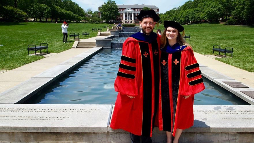 Two PhD students in their graduation regalia, standing in front of the fountain on the central mall of the University campus.