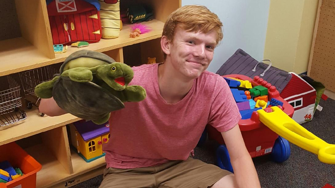 A young man working in a child development lab, sitting on the floor, holding up a large hand puppet of a green frog in his left hand