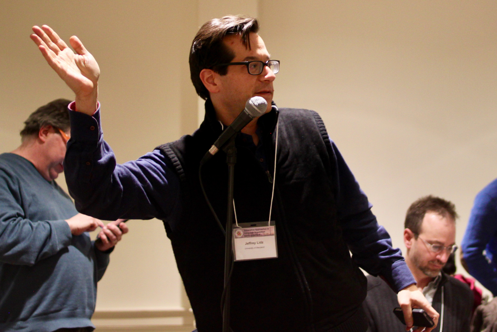 Professor Jeff Lidz, standing at a microphone in profile and talking, with right arm raised in an L, palm up, as if he is about to calmly hurl a ball of invisible energy