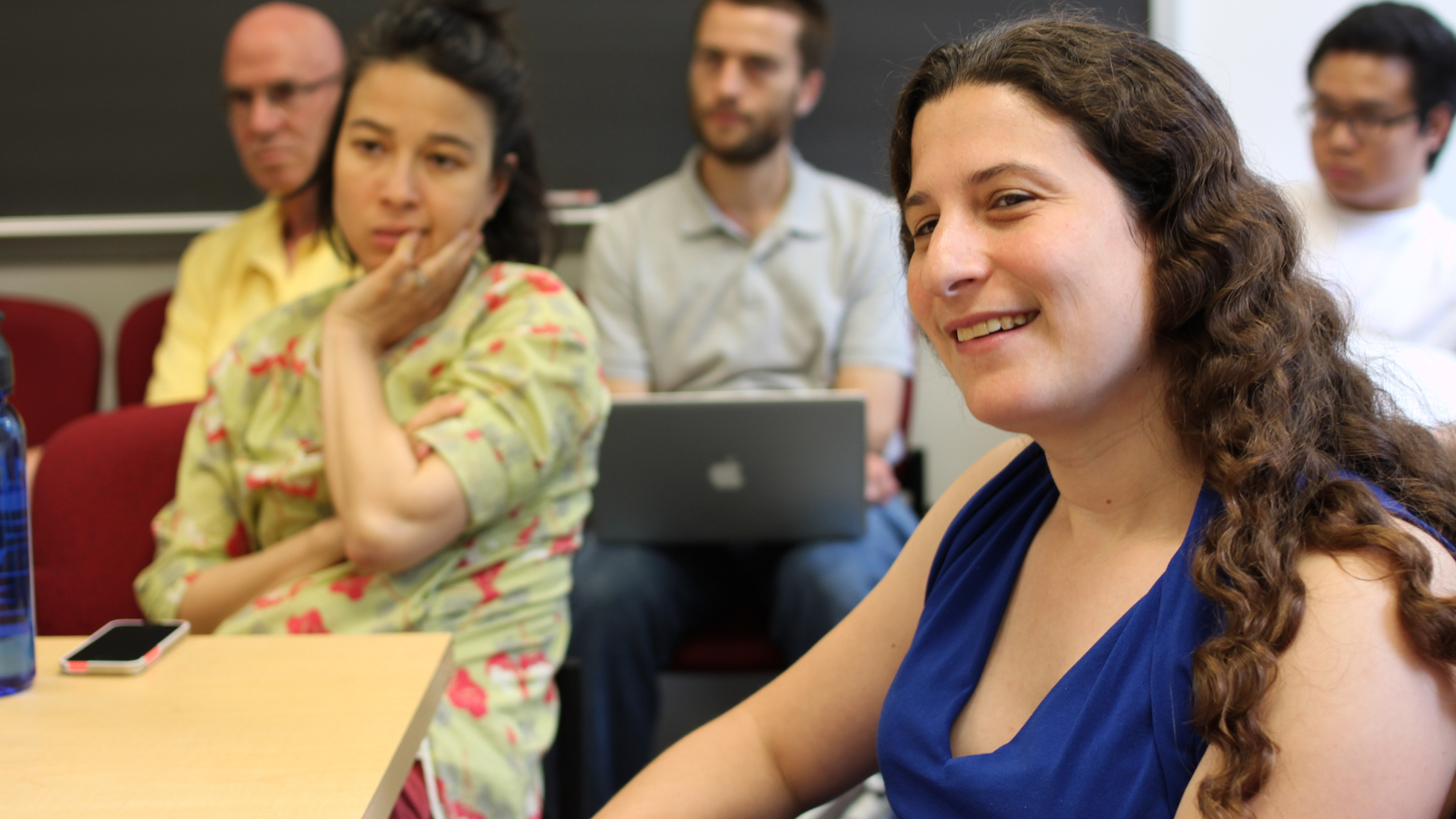 Professor Naomi Feldman in three-quarter profile, smiling, at a dissertation defense, sitting at a conference table with several students and faculty in the background