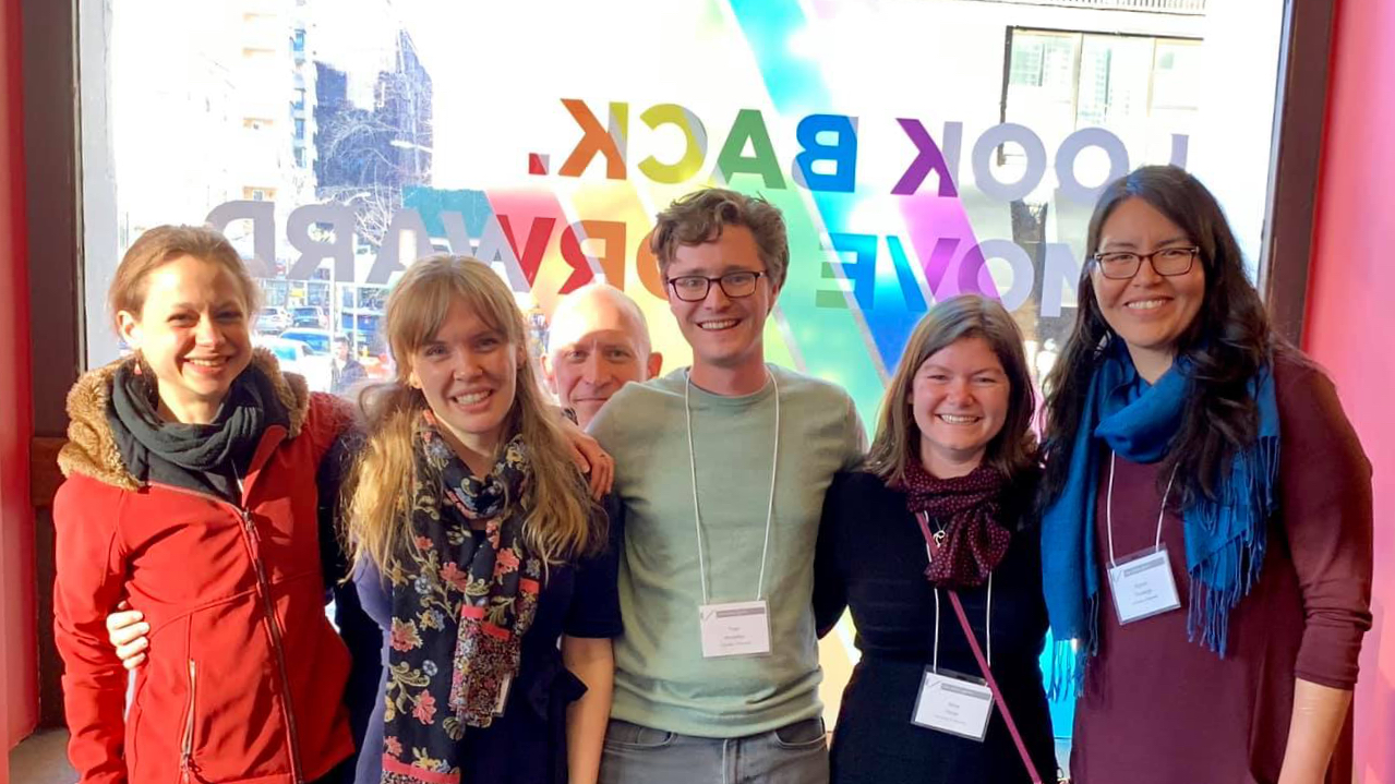 Five PhD students from Linguistics at a conference, standing arm in arm in front of a window