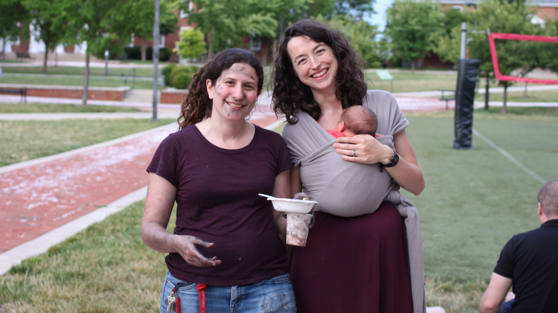 Professors Naomi Feldman and Ellen Lau, smiling at the camera: Naomi is covered in soot from grilling meats in the Argentine style, while Ellen has her newborn son on her chest in a sling