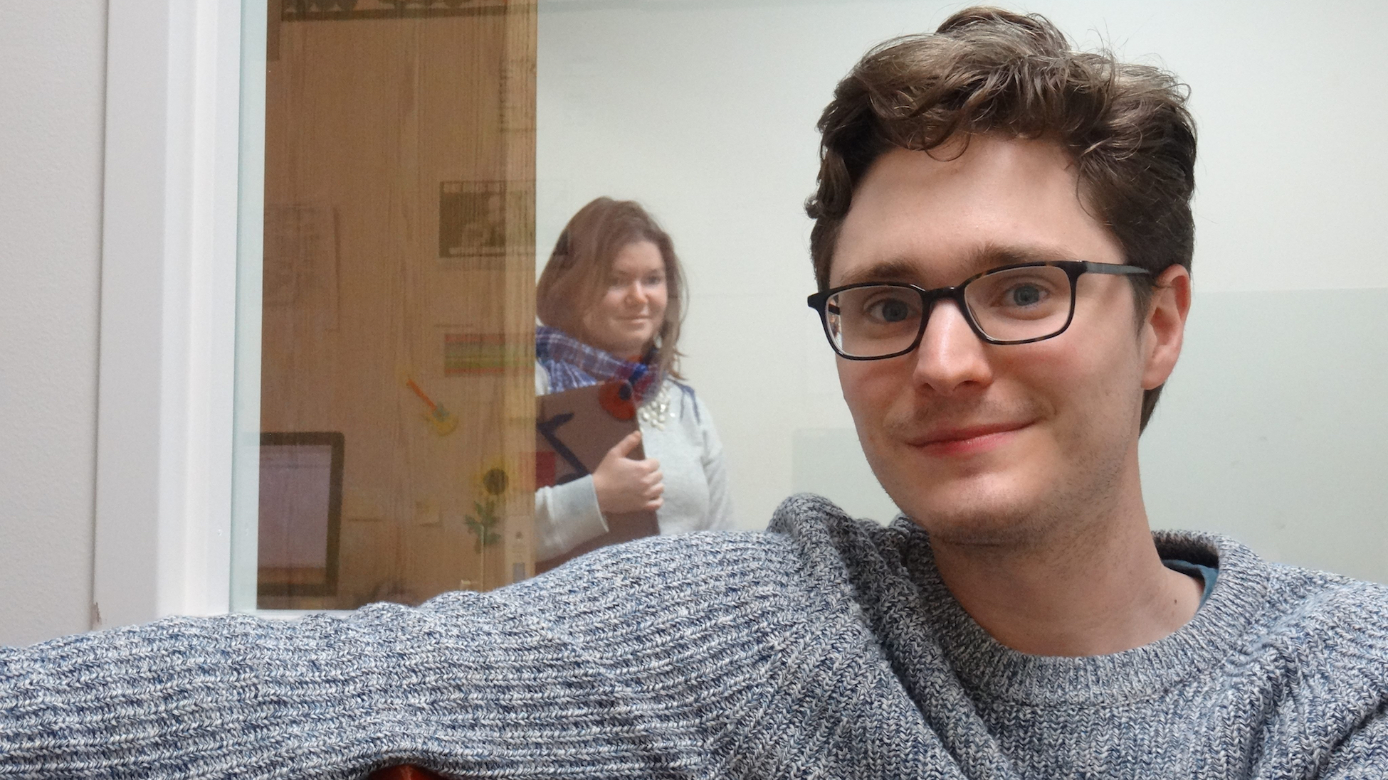 Tyler Knowlton, a PhD student in Linguistics, smiling at the camera, from his office desk, as his office mate skulks in behind him