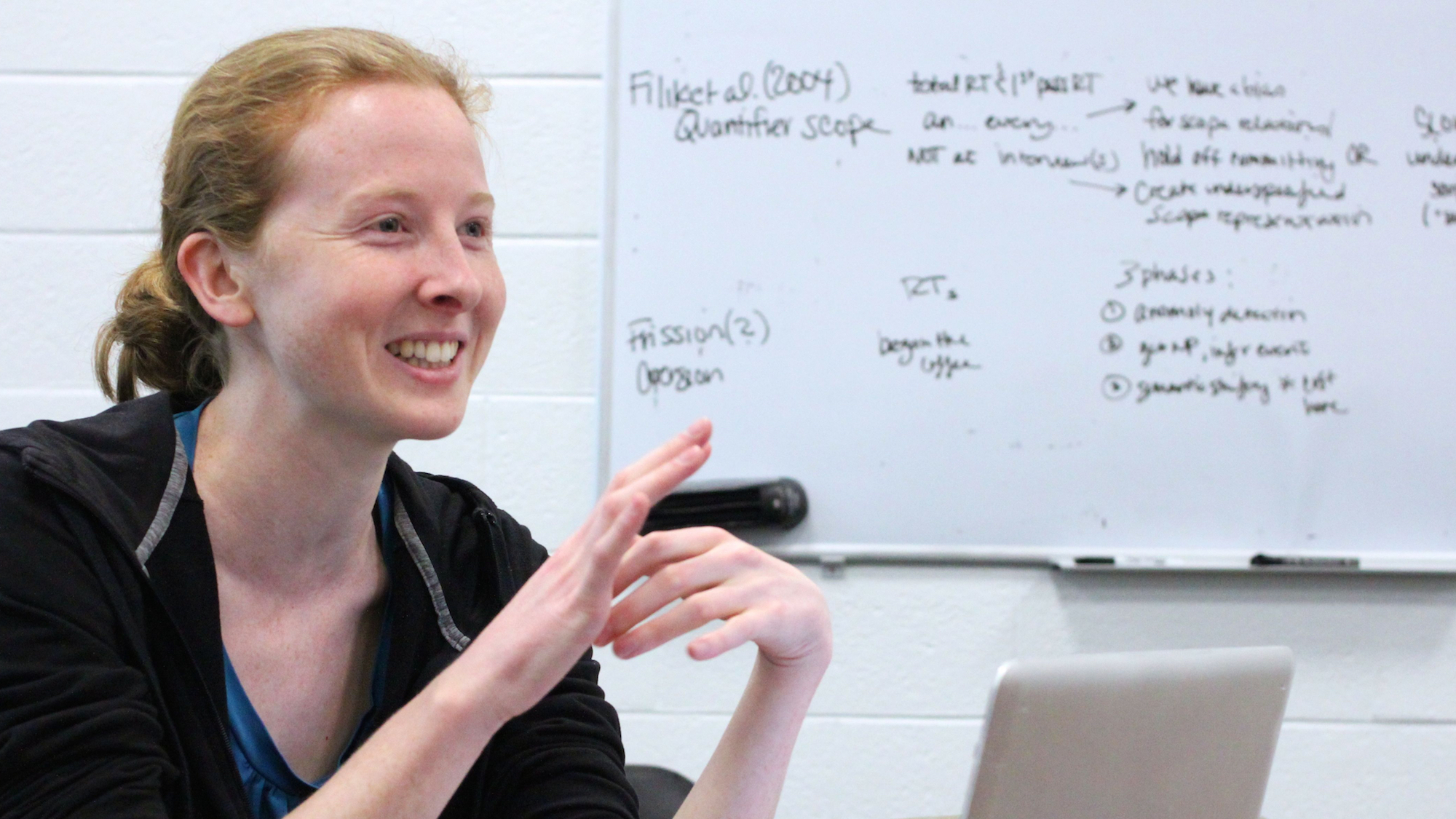PhD student Allyson Ettinger, sitting at a conference table in three-quarter profile, leading a discussion in front of a whiteboard covered in notes, with a gratified smile