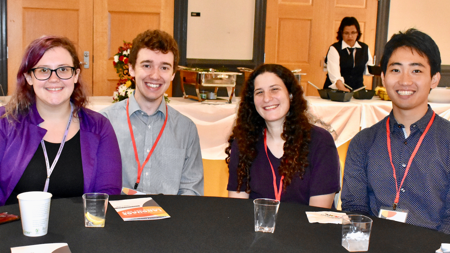 Three PhD students in Linguistics, with a faculty mentor, sitting at a conference, all wearing lanyards for a meeting, smiling at the camera