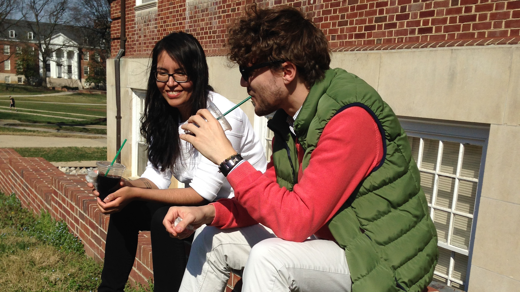 PhD students Sigwan Thivierge and Aaron Dolina, sitting outside on a bright but cool Spring day, wearing light jackets and drinking iced coffees