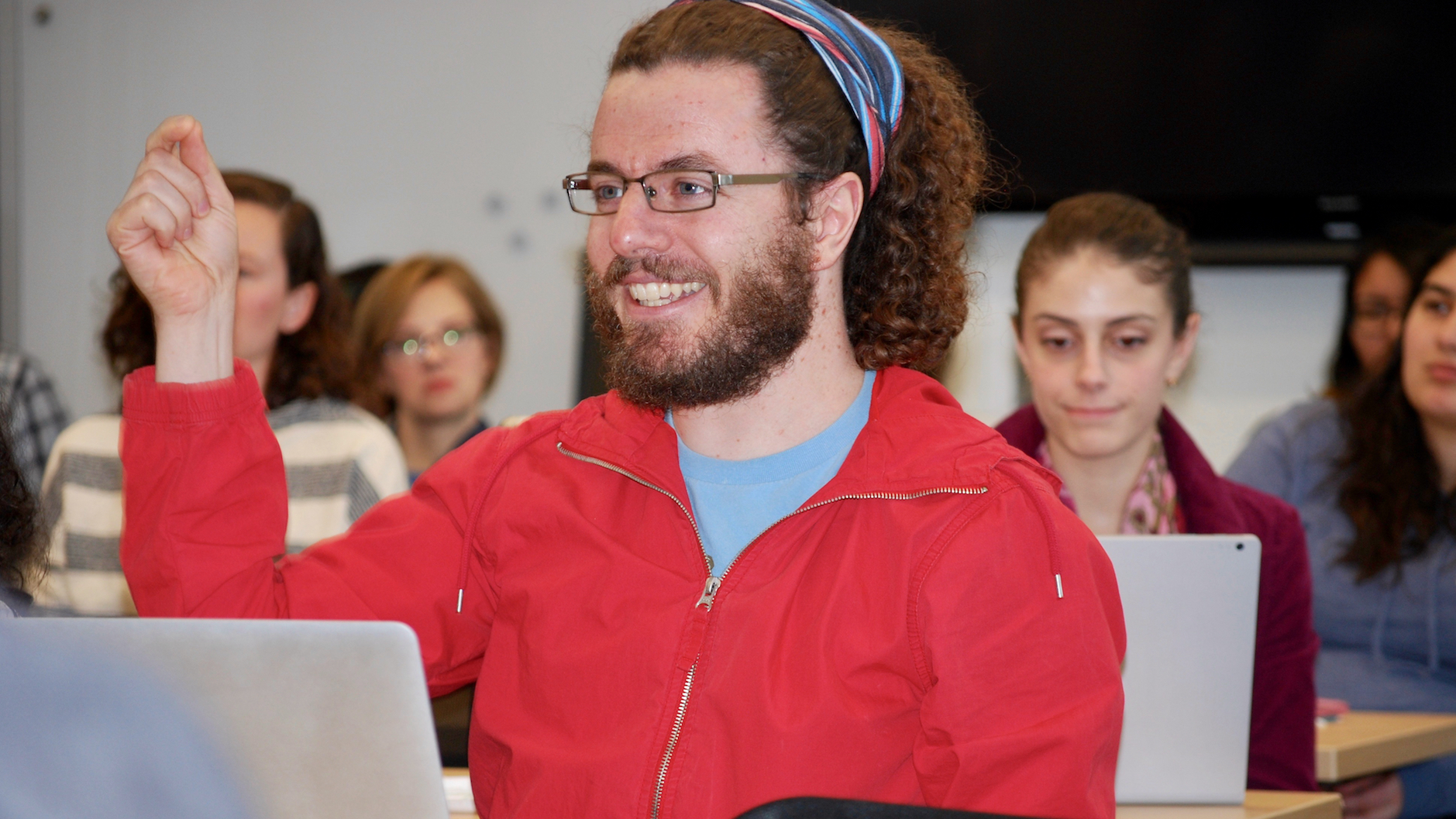 Professor Omer Preminger, seated in three-quarter profile, wearing a red hoodie, smiling as he talks, and pointing up his right hand index finger