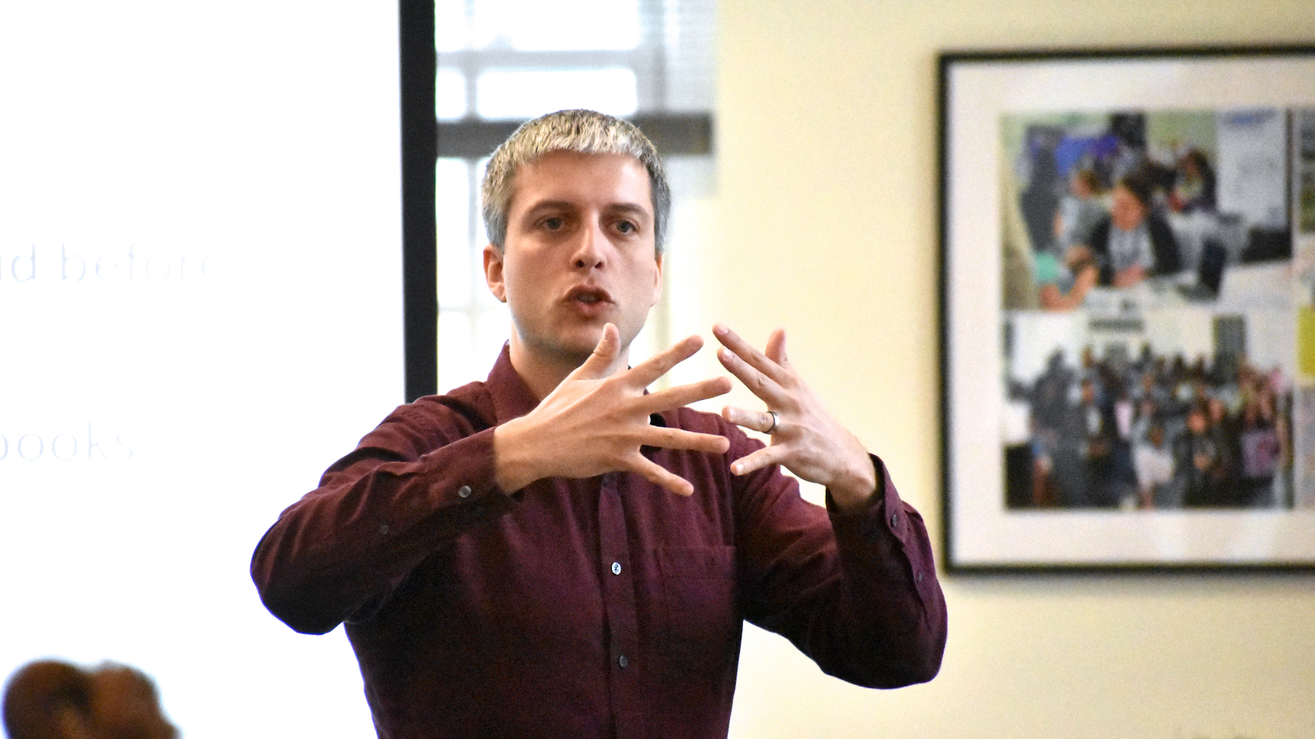 Postdoctoral fellow Dan Goodhue gestures with his hands, almost interdigitating his fingers, will standing up and speaking in a sunlit room