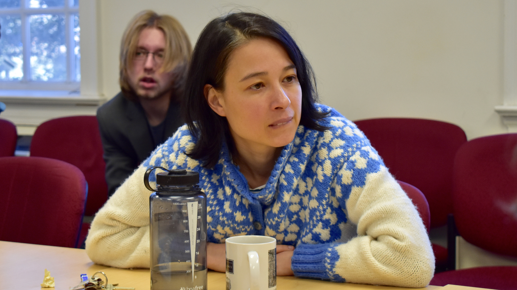 Professor Valentine Hacquard in a ski sweater, arms resting folded on a conference room table, looking off to the right