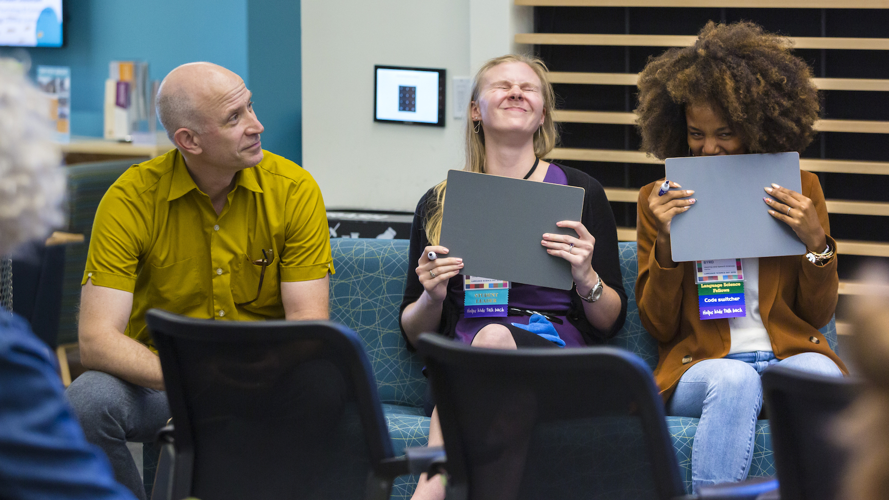 A panel of three people, seated. One of them, Professor Alexander Williams, looks rightward at two young female students, who are convulsed with laughter