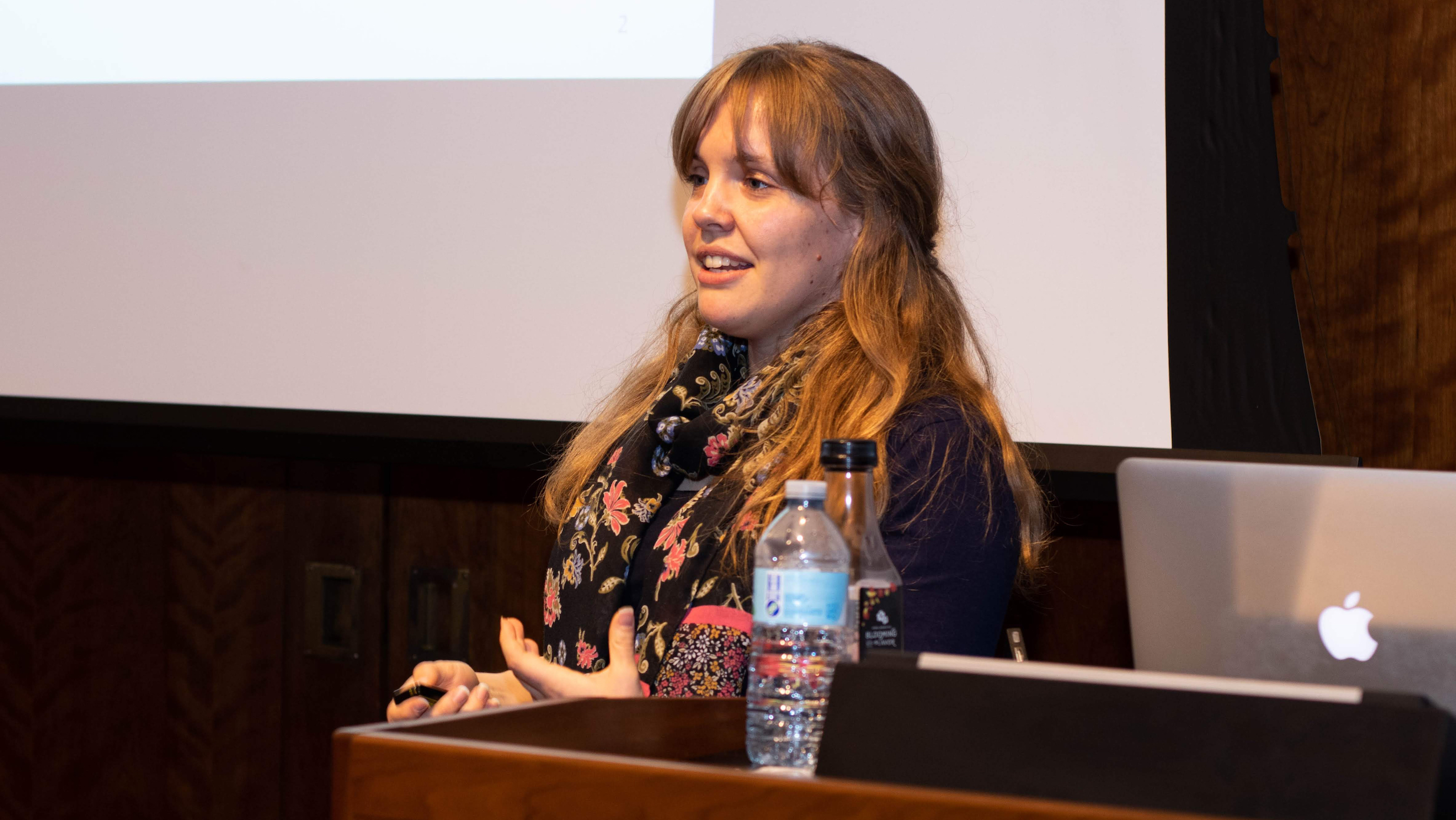 PhD student Annemarie van Dooren, sitting at a table in three-quarter profile, in front of a large screen at the front of a lecture hall, leading a lecture