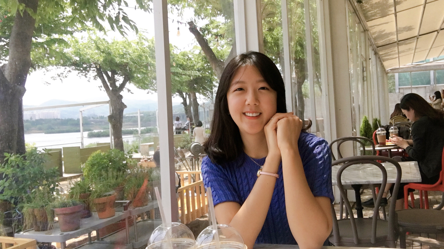 PhD student Eun-Kyoung Rosa Lee, smiling at a table in a sunlit cafe, elbows on the table and clasped hands near her cheek, with two cups of bubble tea in the foreground, and green trees visible behind her through large windows