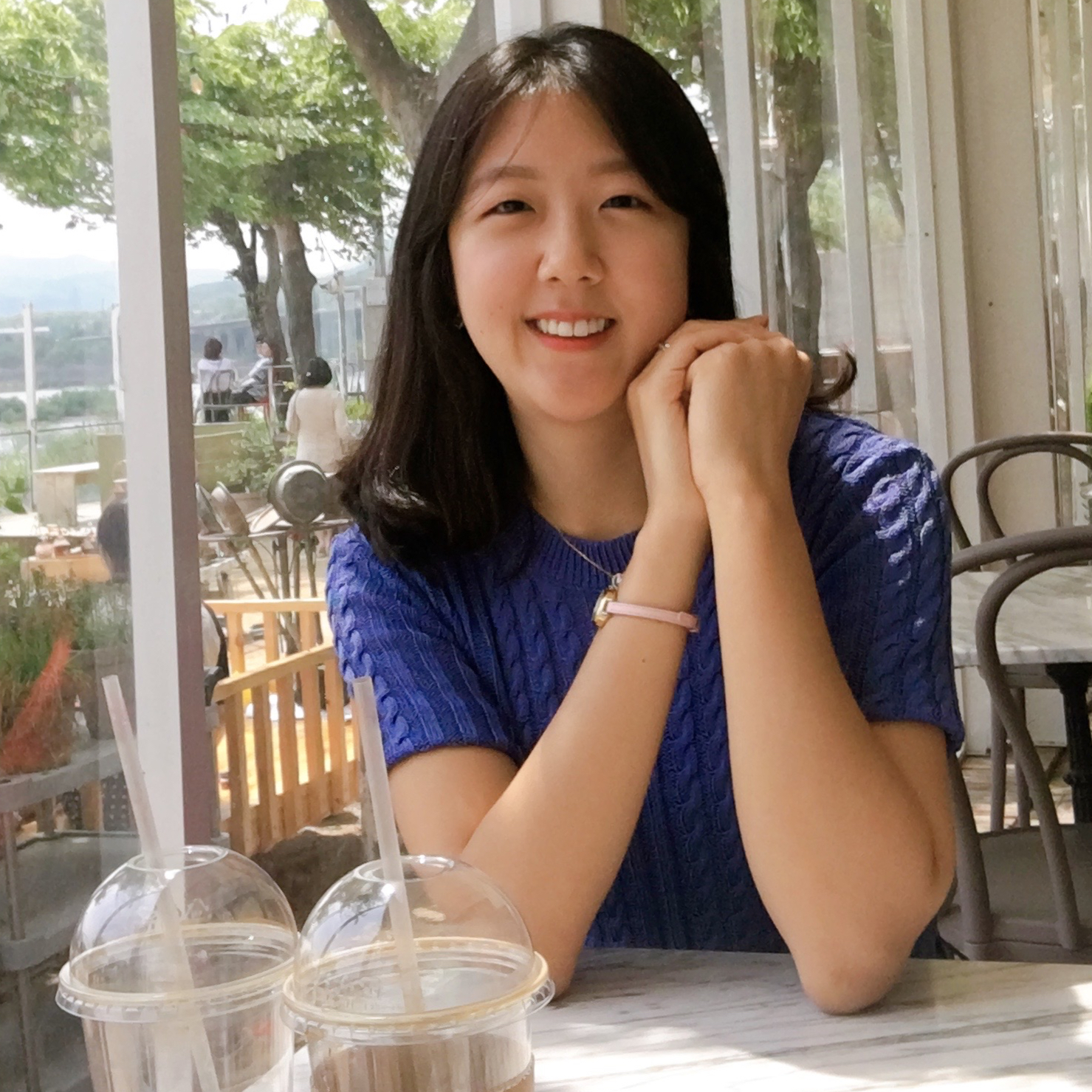 PhD student Eun-Kyoung Rosa Lee, sitting at a table in a sunlit cafe, trees visible through large windows behind her, and two cups of bubble tea in the foreground