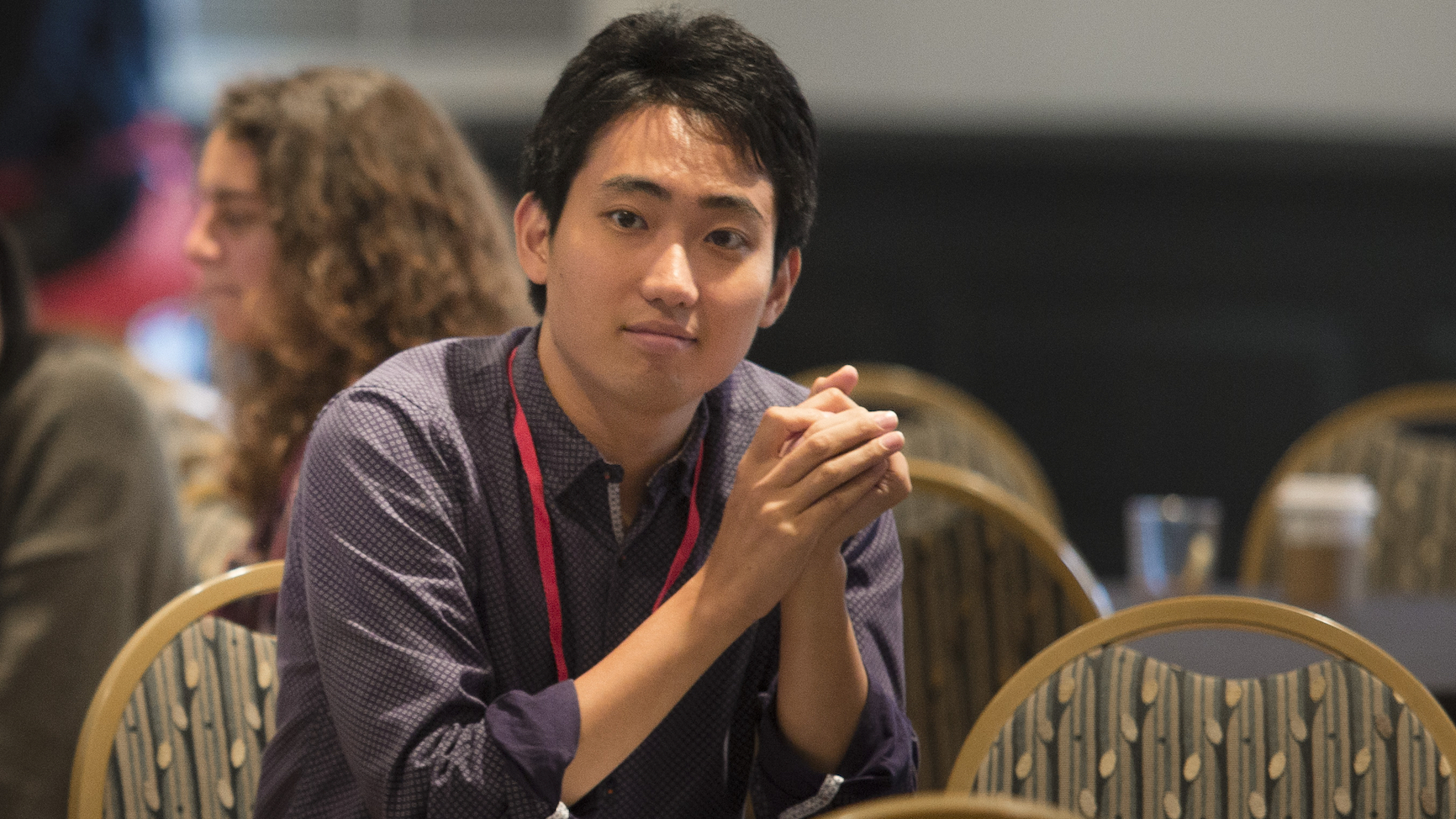 PhD student Masato Nakamura sitting at a table, resting on his elbows and hands clasped in front of him, grinning
