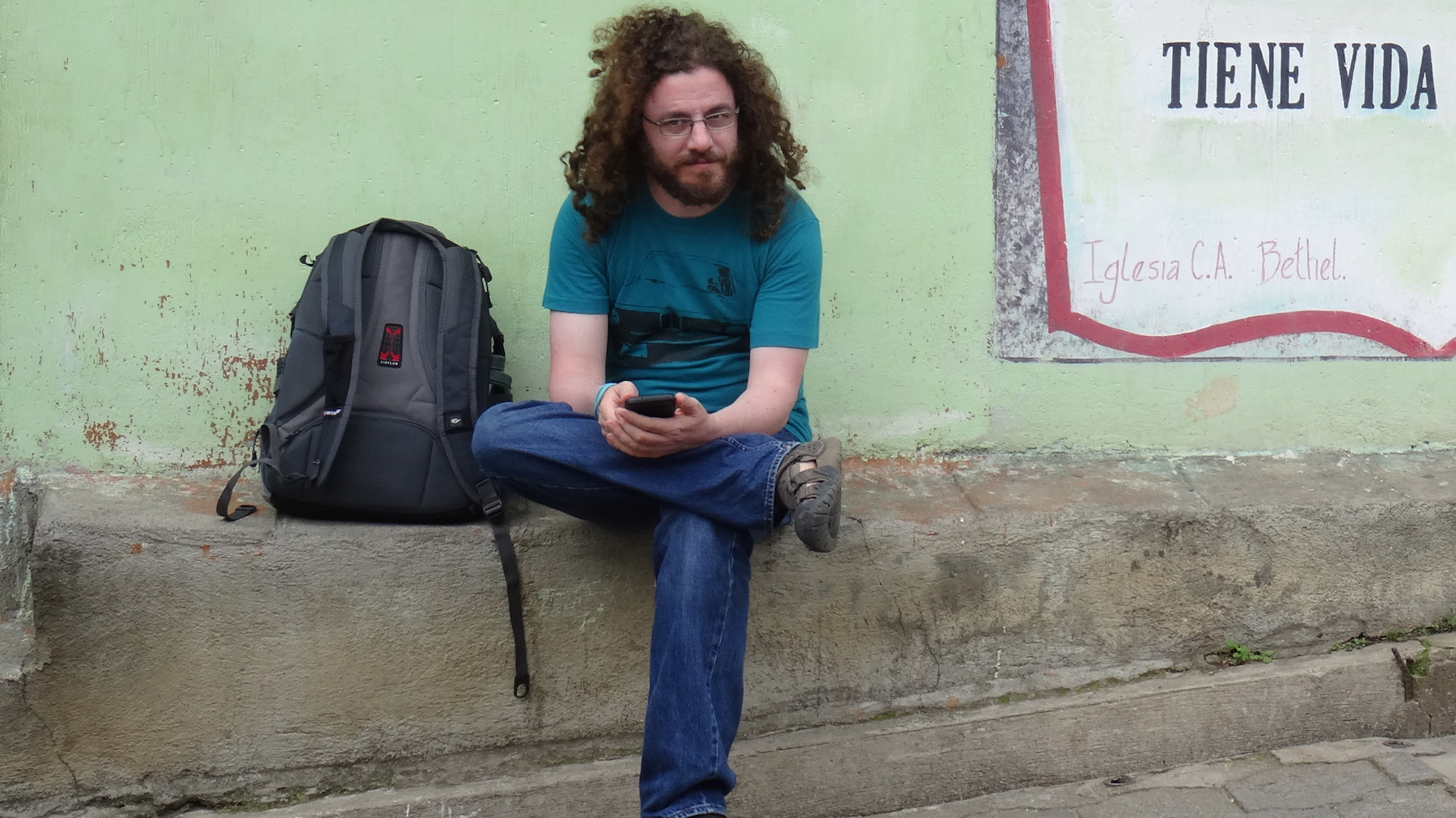 Professor Omer Preminger, sitting on a street in Guatemala, in front of a minty green wall, checking his phone