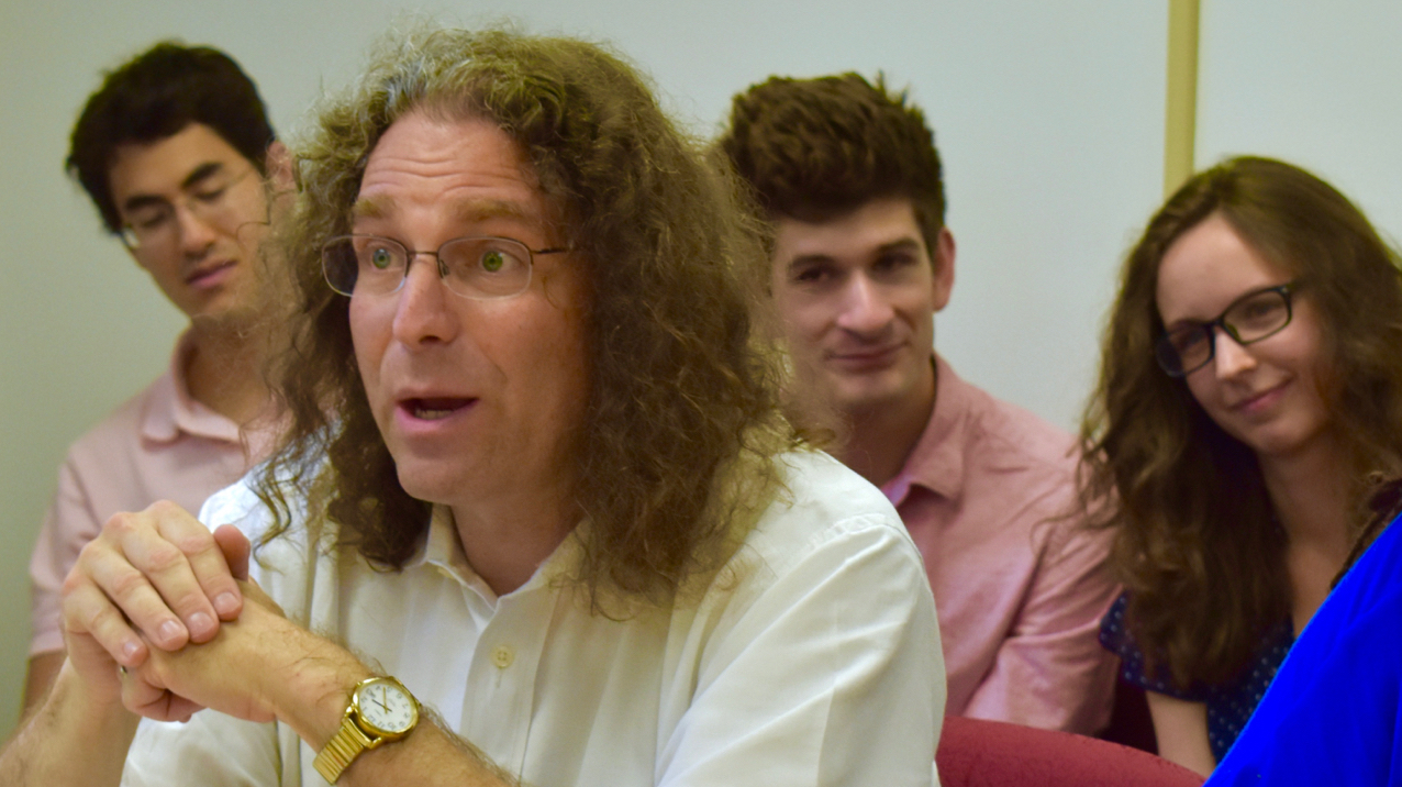 Professor Philip Resnik in three-quarters profile, asking a question at a dissertation defense, with three students behind him, smiling.