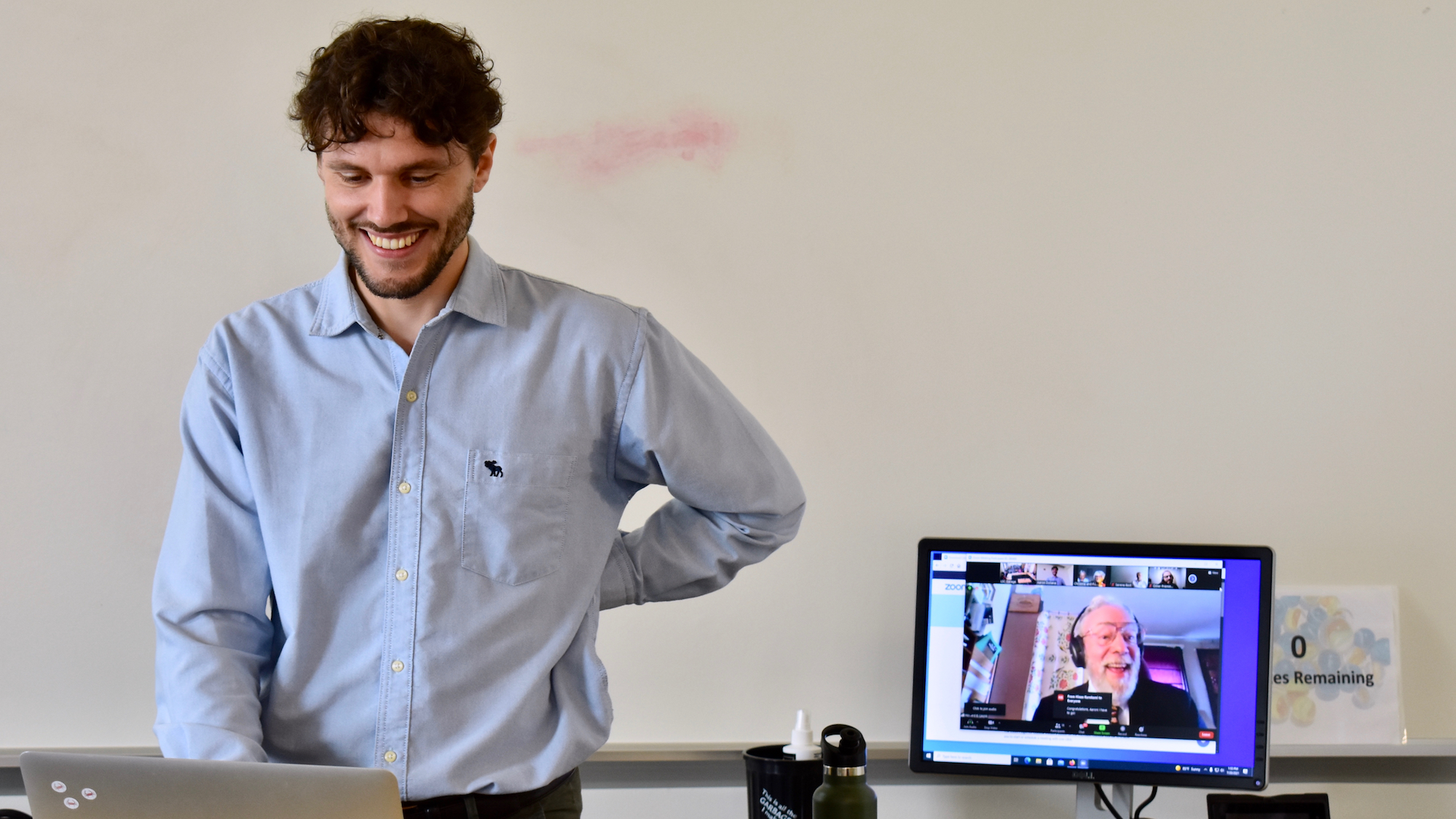 PhD student Aaron Doliana stands at the left, looking down at a laptop screen, smiling, while behind him on the right, another computer displays what Aaron sees, his advisor Howard Lasnik smiling back.