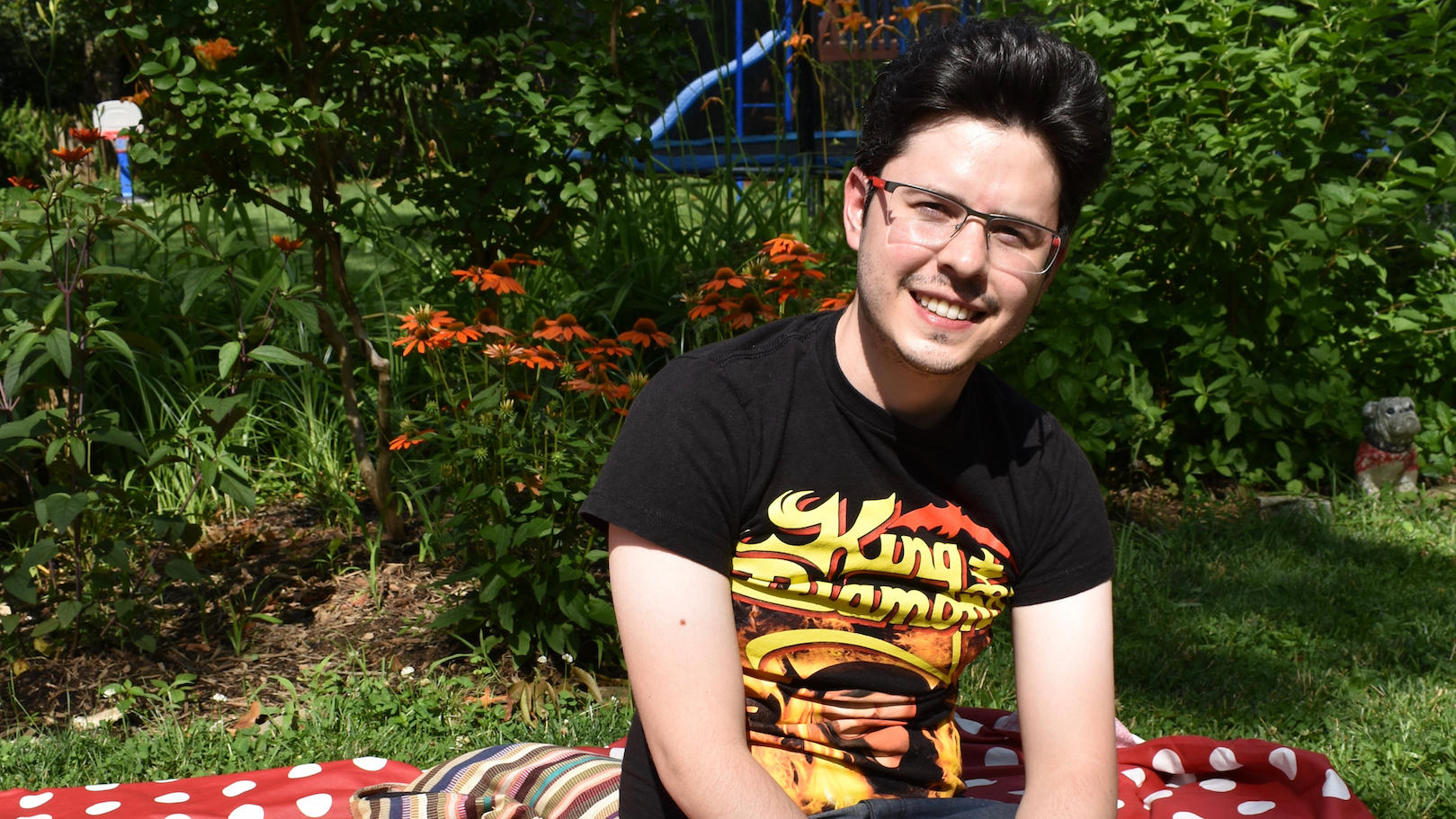 PhD student Rodrigo Ranero, sitting in a garden under summer sun, orange echinaceas in the background, wearing a "King Diamond" t-shirt.