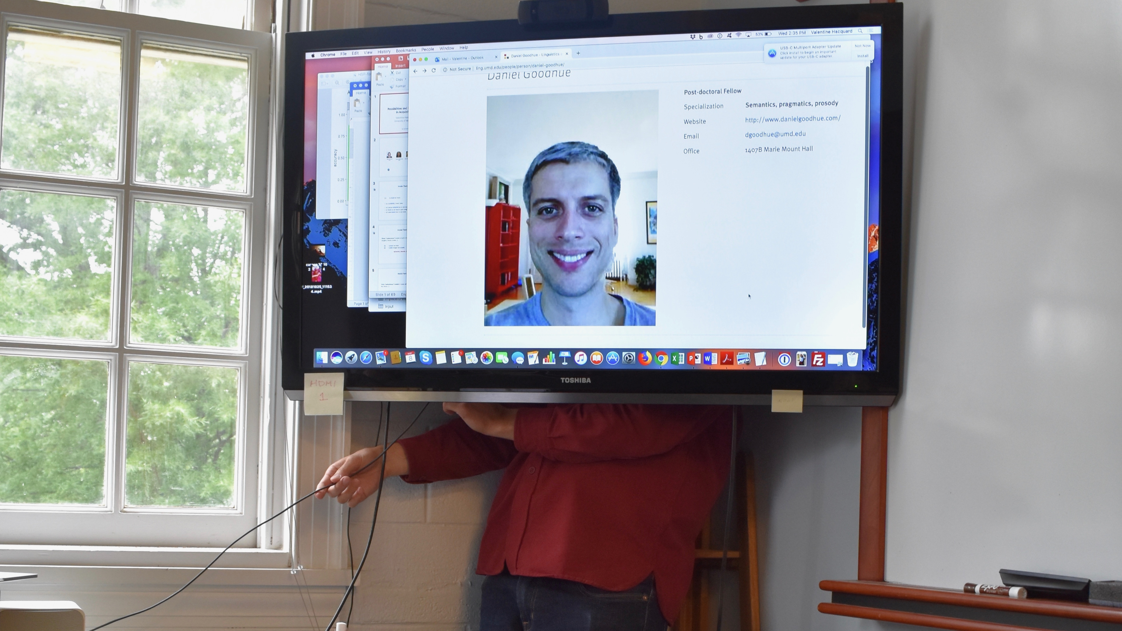 A man standing behind a TV screen, fixing the wires, as the screen displays a portrait of that man's head