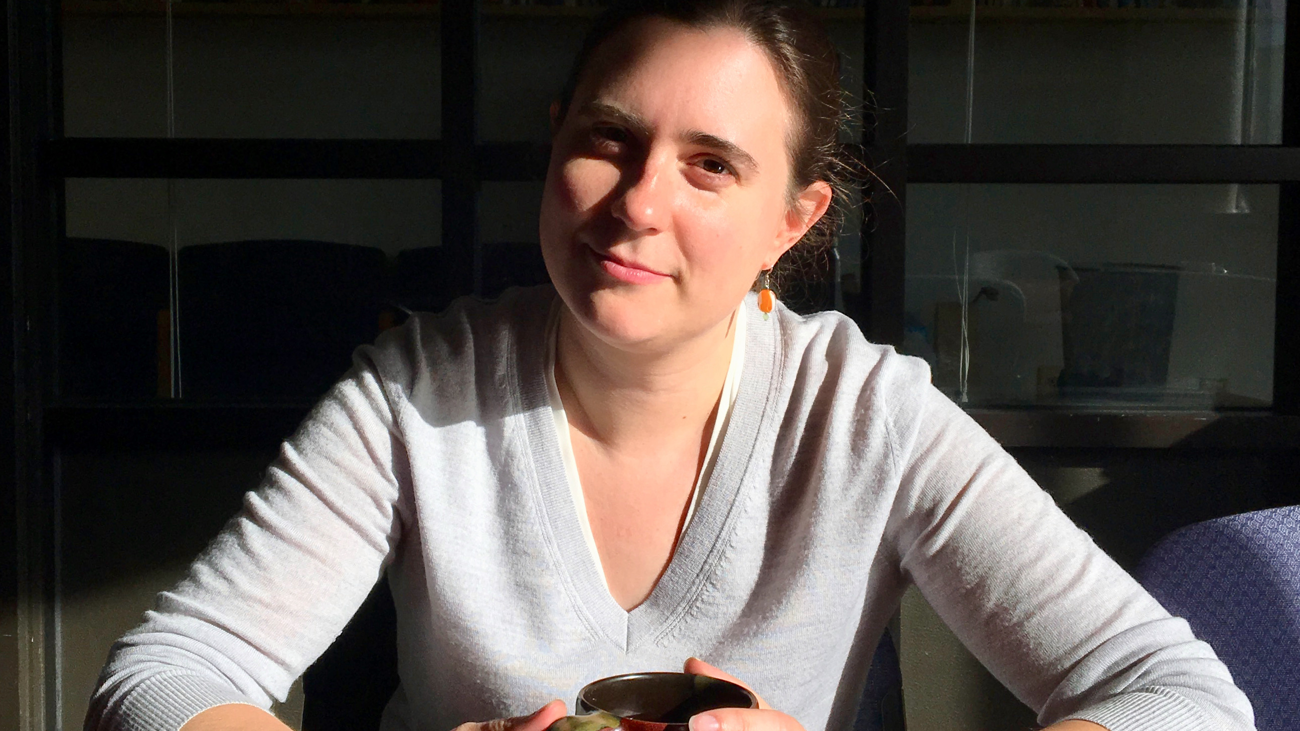 PhD student Laurel Perkins, facing the camera with a determined expression, sitting at a table in heavy shadow, holding a cup of tea in front of her