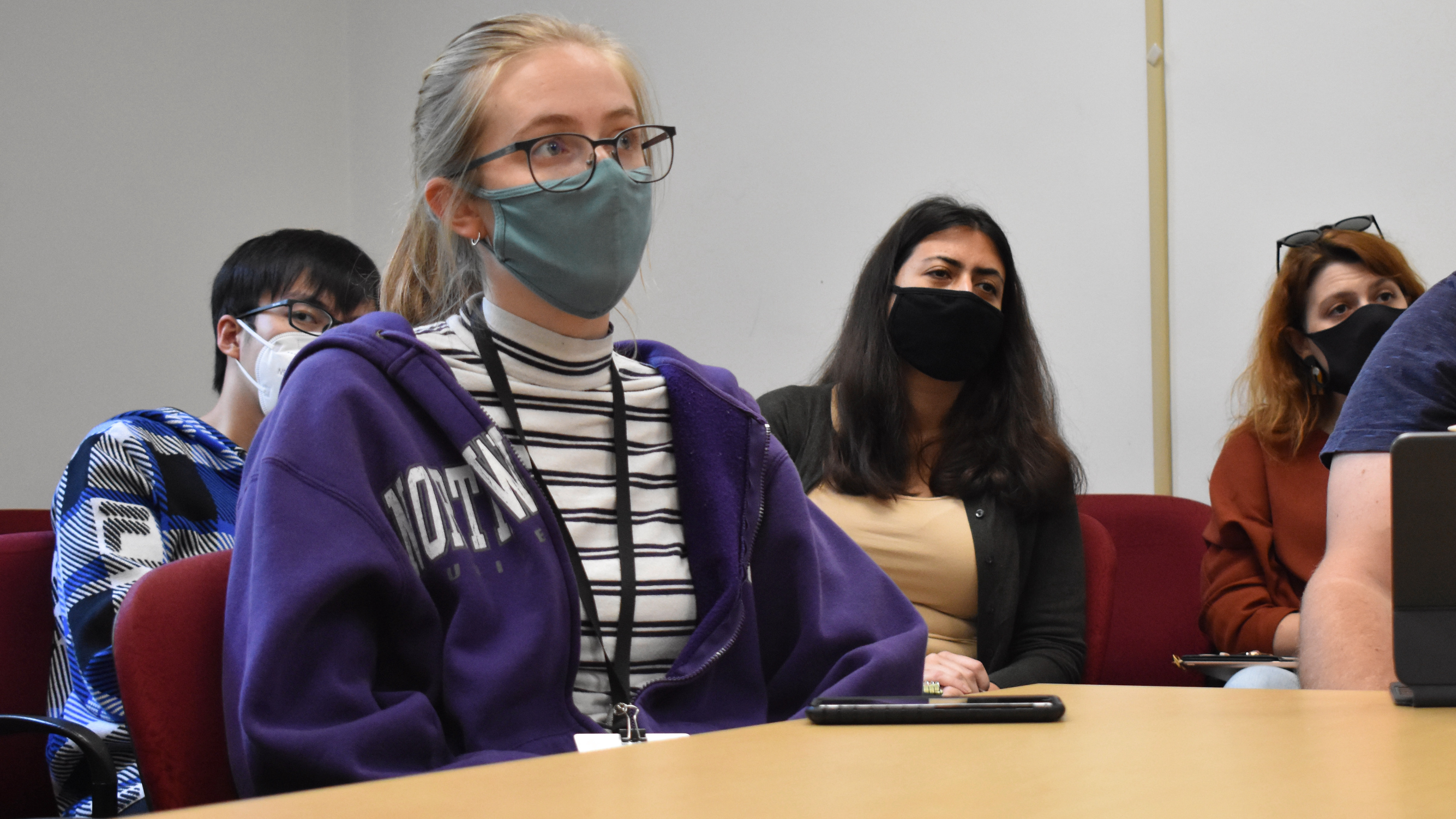 A young woman in a purple sweatshirt and a covid facemask, sitting at a table and listening intently to a seminar discussion