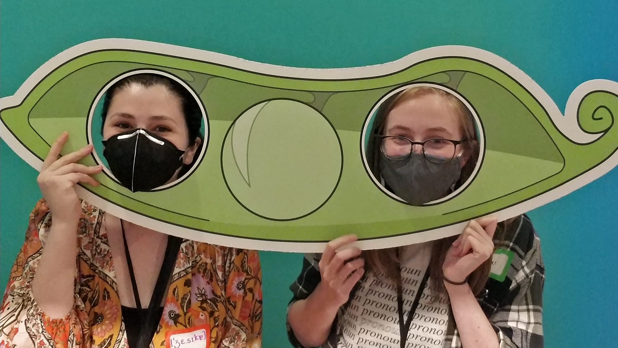 Two young women, their faces showing through holes in a giant cut out of a bean, as if they were two peas in a pod.
