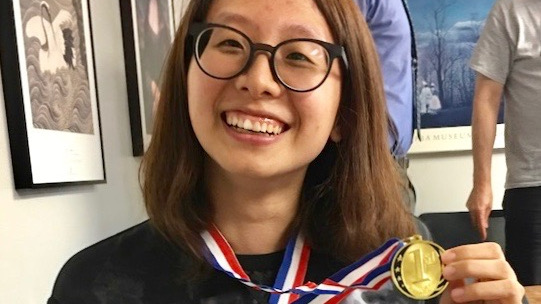 A close portrait of the smiling face of Yu'an Yang, a PhD student in linguistics, holding up a gold medal that hangs from her neck by a ribbon