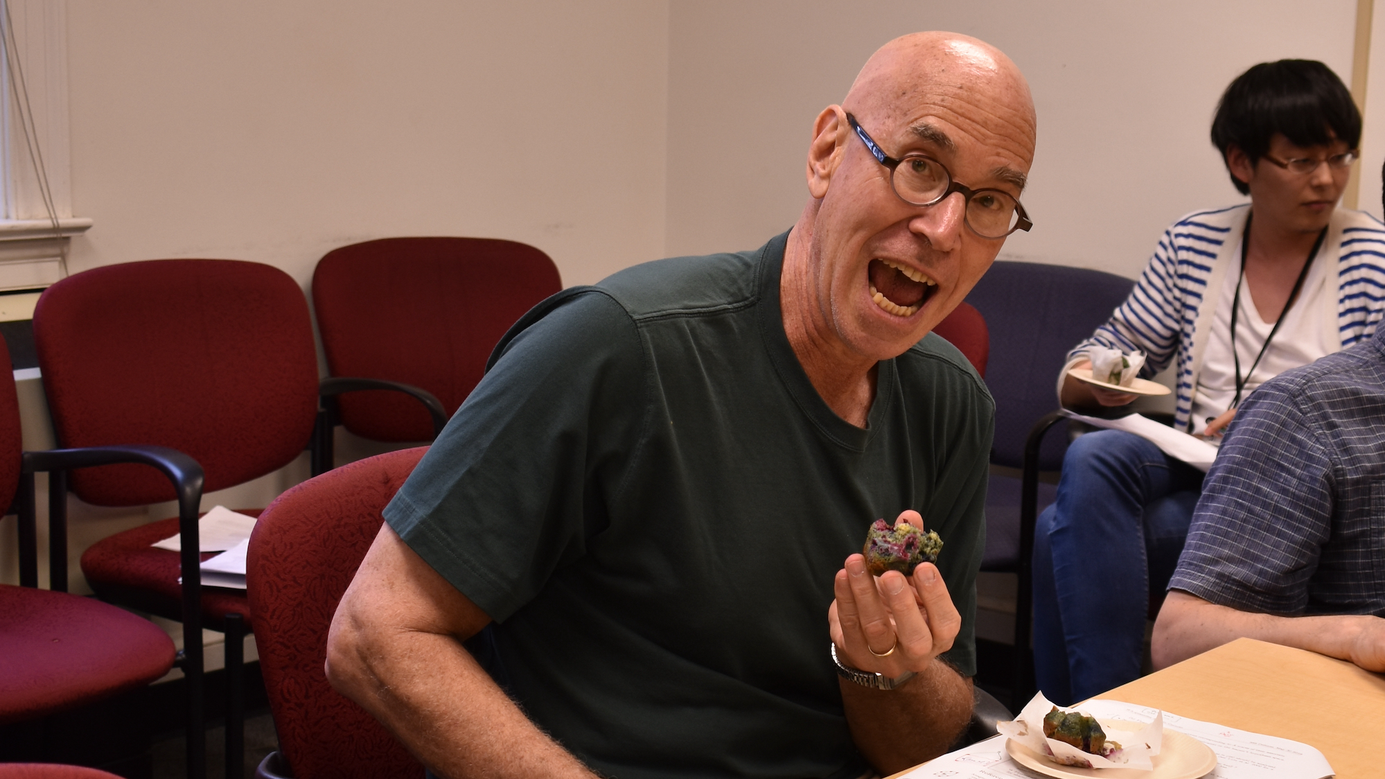 A bald man in a green polo shirt, leaning dramatically towards a cupcake in his left hand, mouth wide open and mugging for the camera, pretending to plan to wolf it down whole