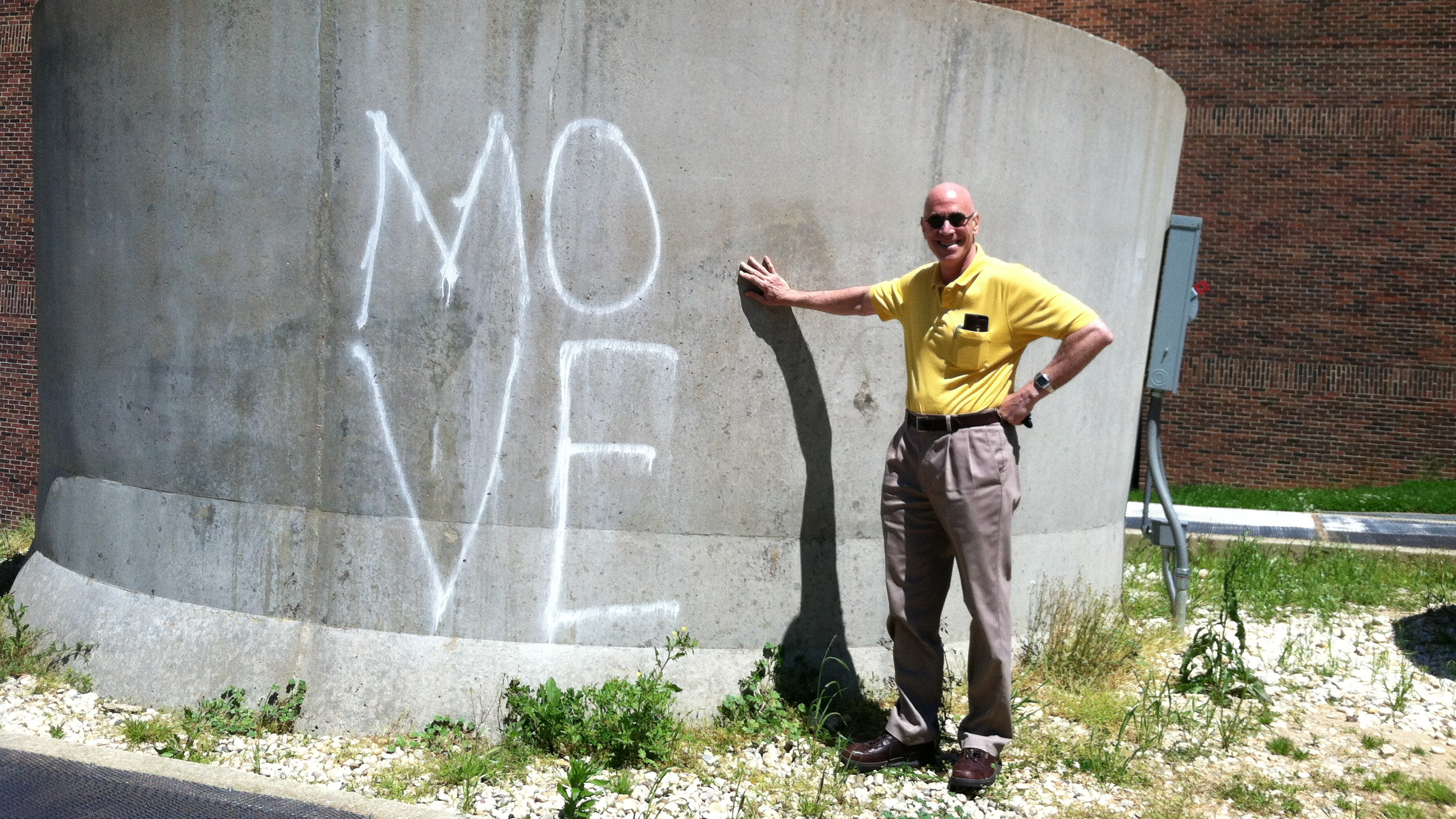 A bald man in a yellow polo shirt, standing next to an HVAC chimney, onto which someone has spray-painted the word "move".
