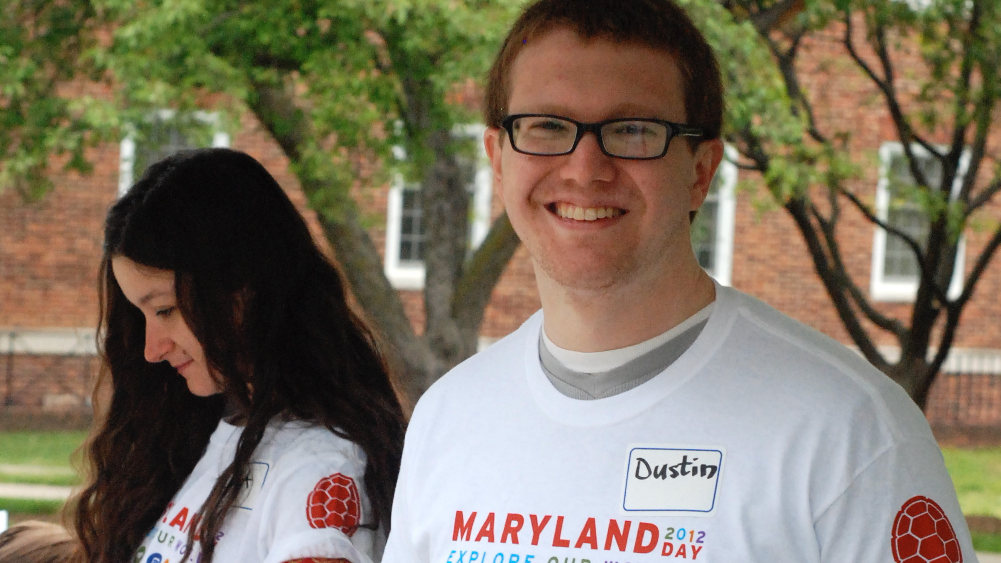A young man in glasses standing outside, wearing a "Maryland Day" t-shirt, smiling at the camera.