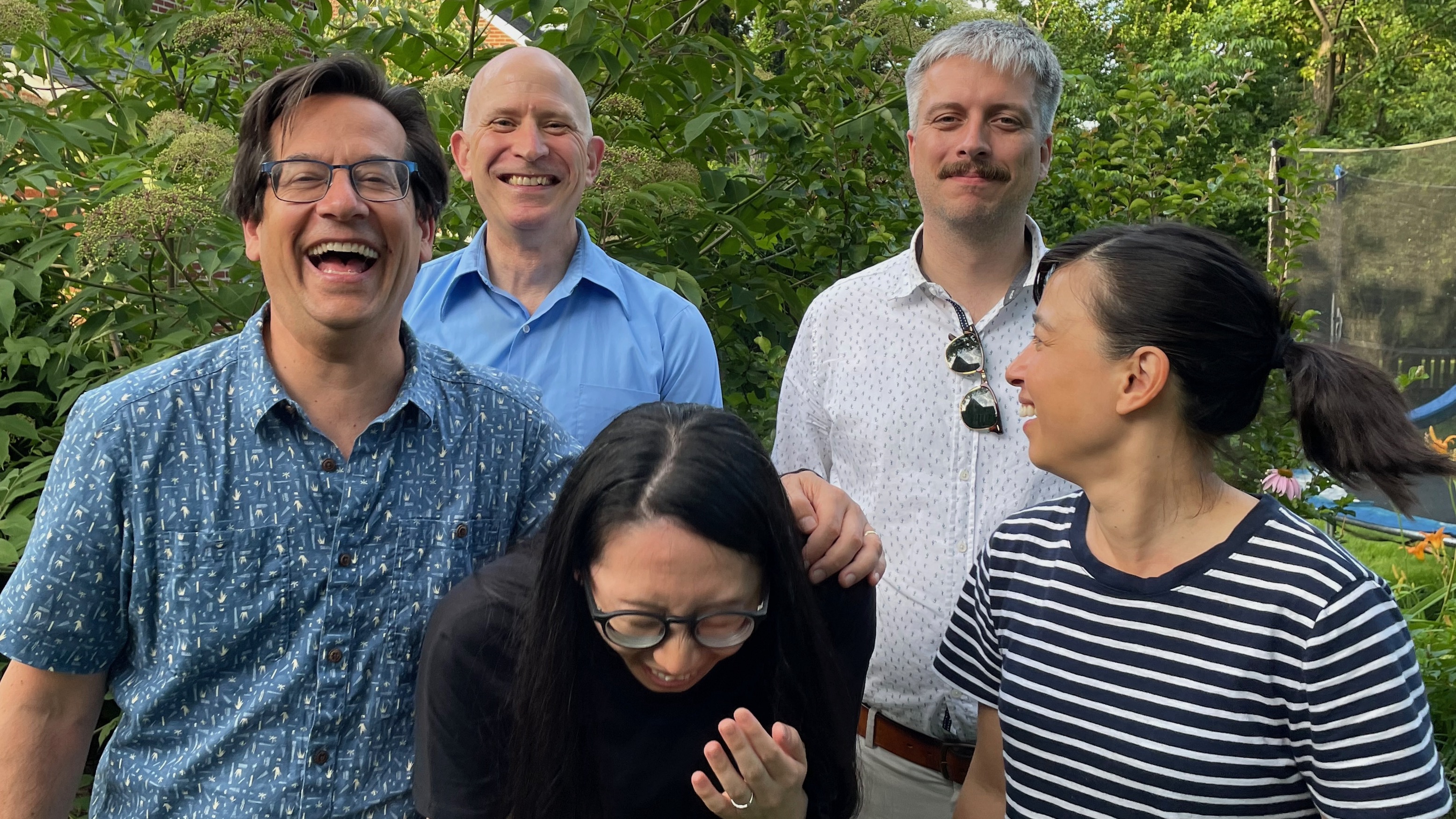 A group of five people in a garden, grouped for a photo, bursting into laughter.