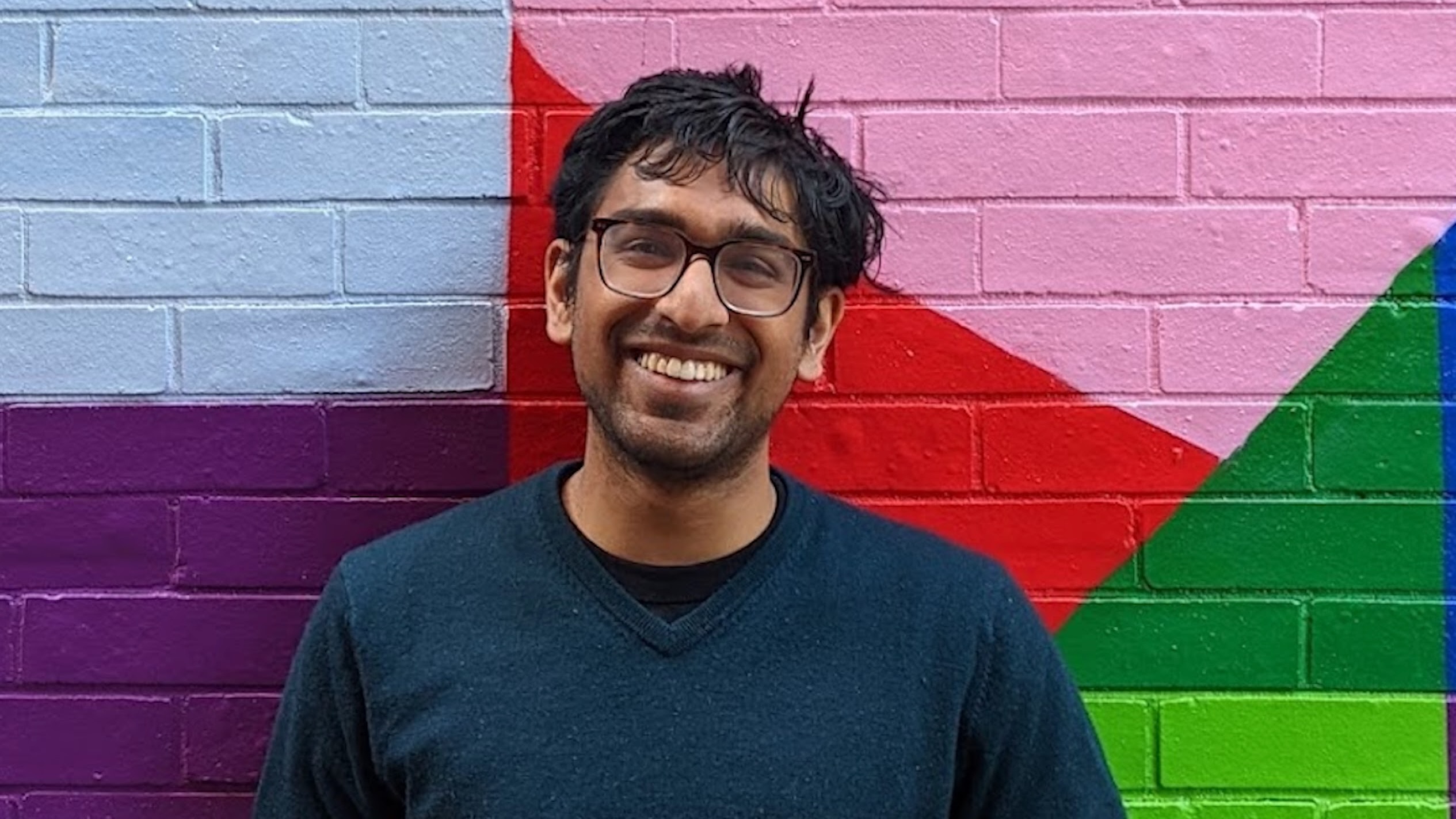 Sathvik Nair, PhD student in Linguistics, standing in front of a brick wall painted colorfully with geometric shapes, smiling broadly.