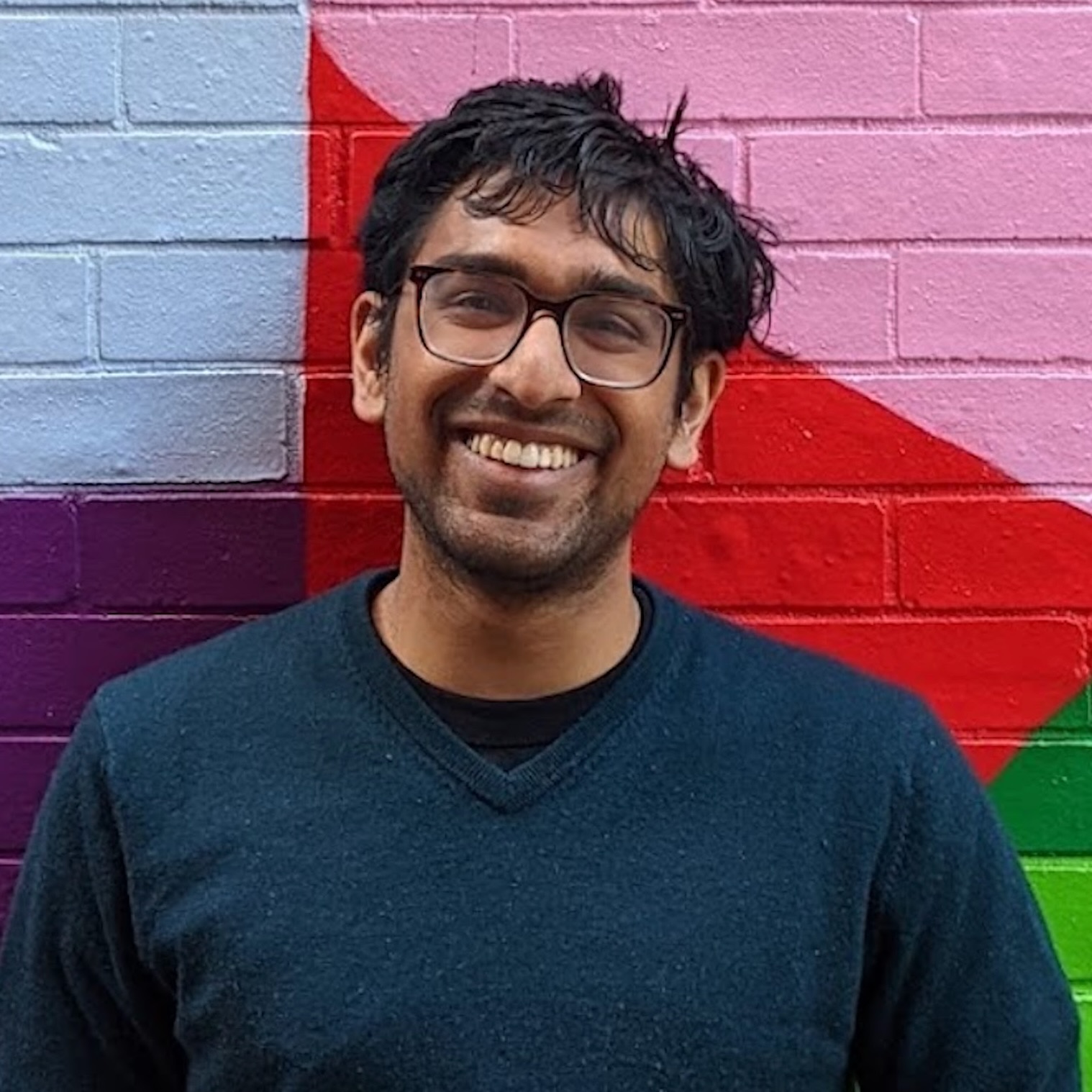 Sathvik Nair, PhD student in Linguistics, standing in front of a brick wall painted colorfully with geometric shapes, smiling broadly.