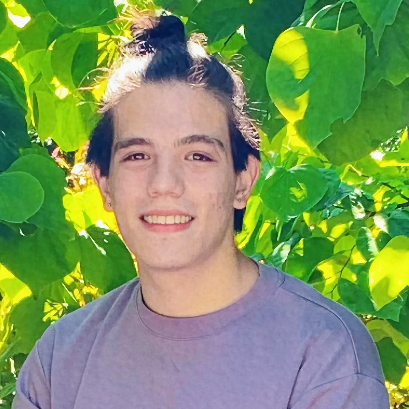 Sebastián Mancha, PhD student in Linguistics, smiling at the camera, standing in front of the bright green leaves of a tree in a DC garden.