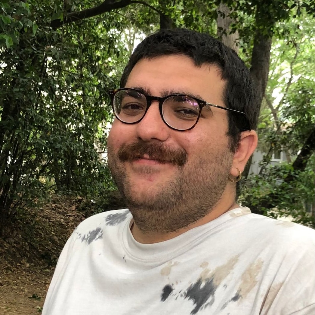 Portrait of Utku Turk, PhD student in Linguistics, a young man with glasses and a mustache, smiling at the camera, sitting in front of some trees.