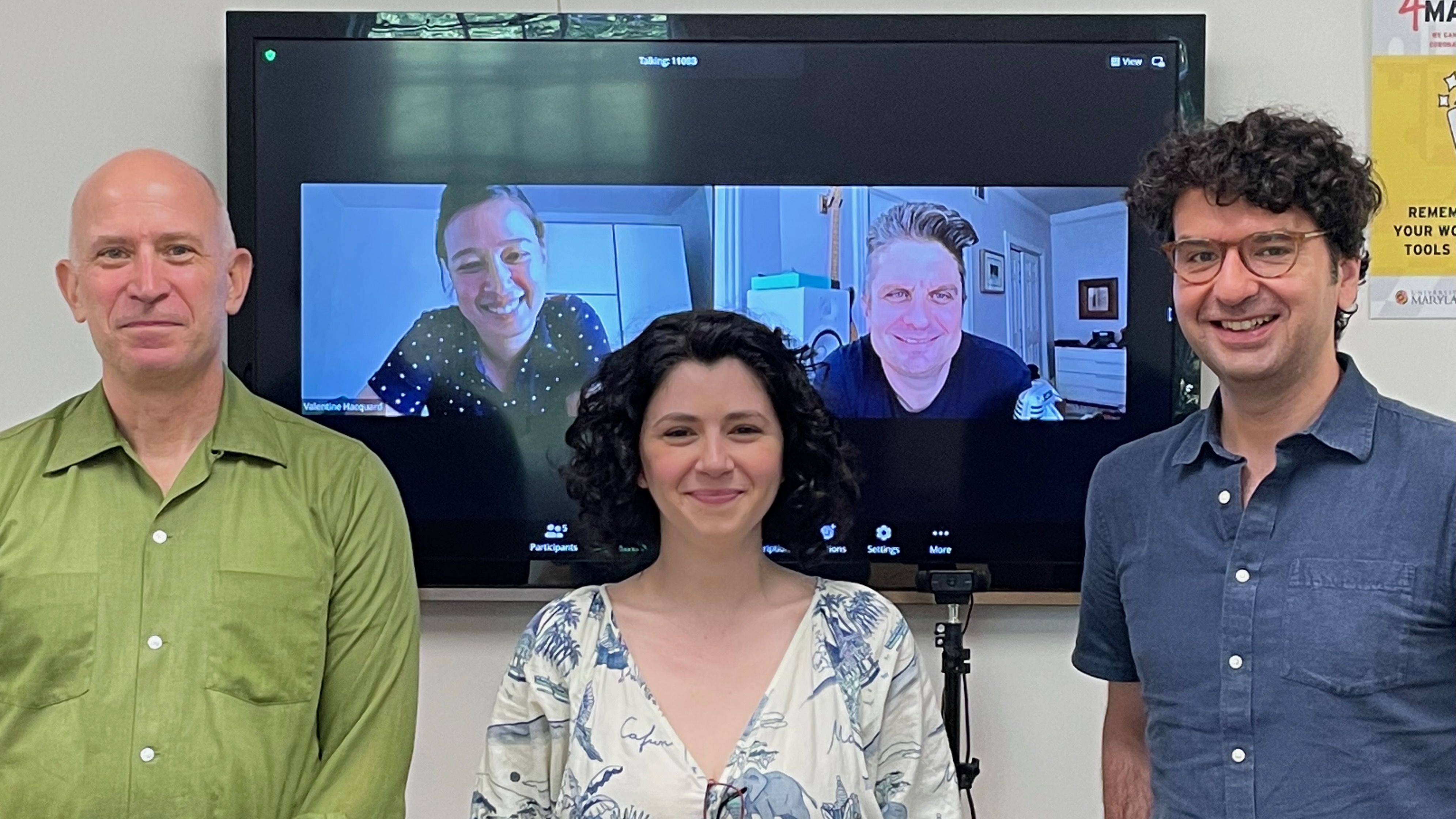 A young woman, smiling after having defended her qualifying paper, flanked by her proud faculty committee members: two men standing on either side, and a man and a woman joining remotely on a large screen behind her.