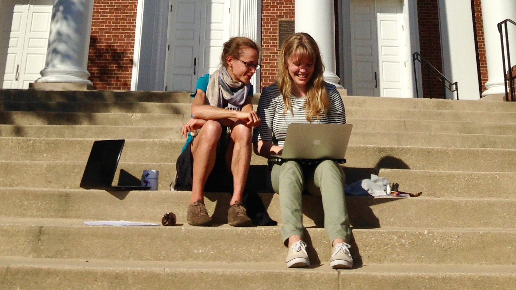Two young women sitting on the steps of Memorial Chapel, one looking over the shoulder of the other at her laptop.