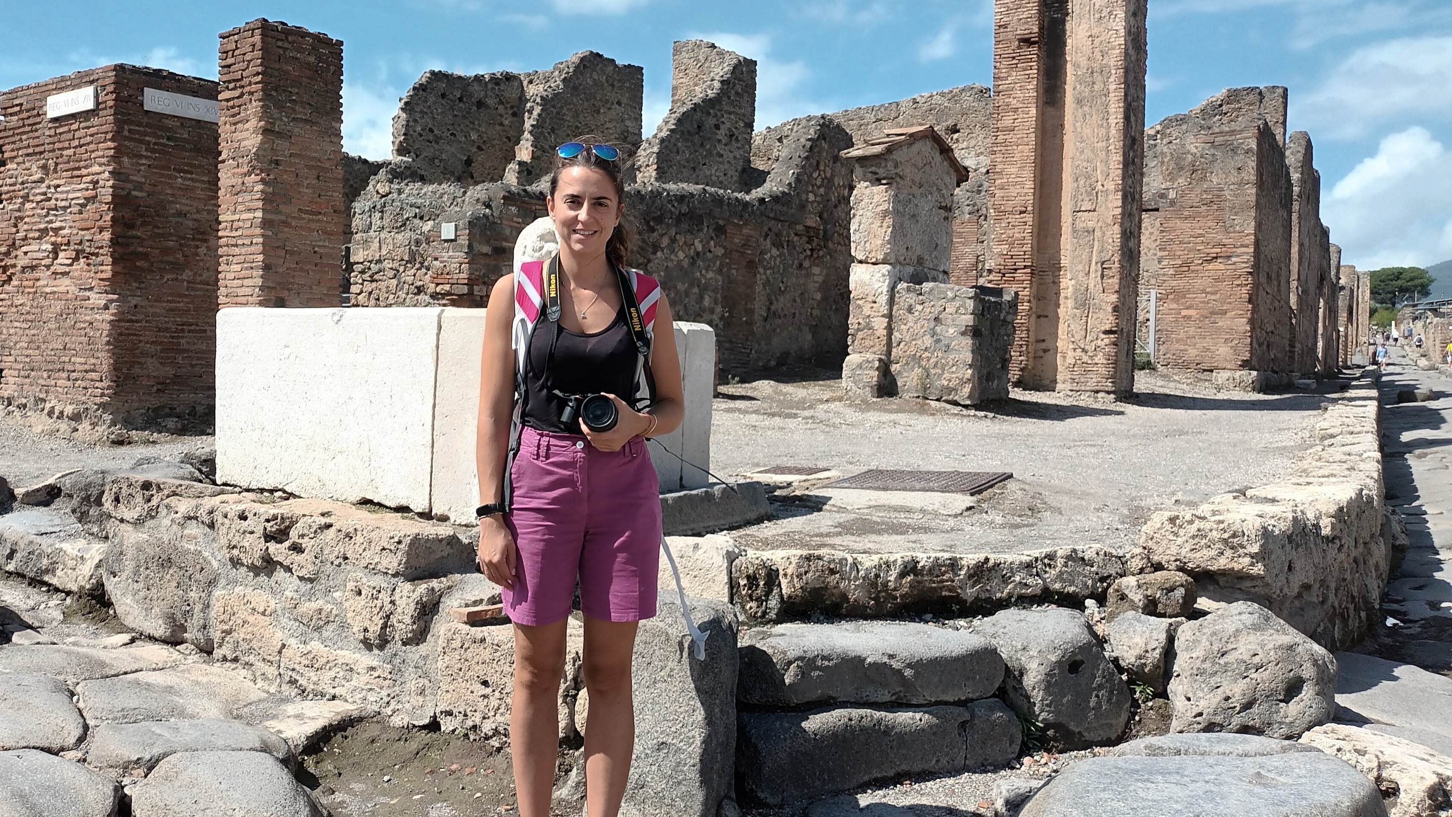 Luisa Seguin, PhD student in Linguistics, standing in front of the ruins of Pompei, in raspberry shorts and a black tank-top, holding a camera in her left hand and smiling.