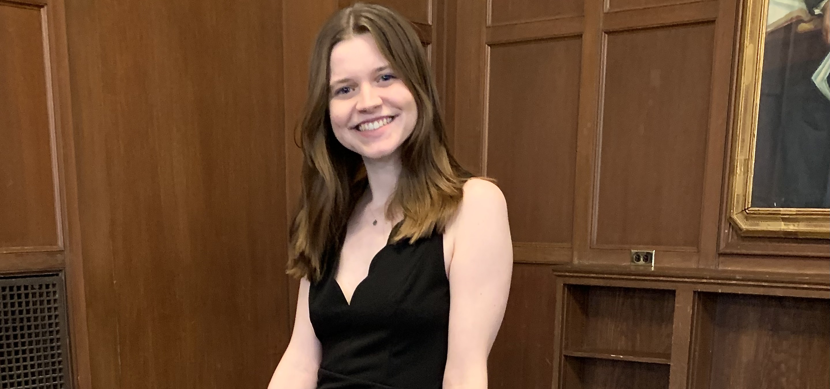 A young woman in a black dress, sitting in a wooden paneled room in three-quarter profile, smiling broadly.