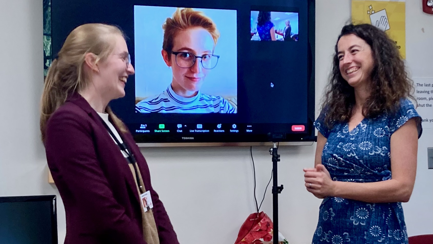 A tv screen displaying a portrait of a young woman, that same woman standing to the left of it, receiving the congratulations of her advisor, Professor Ellen Lau, who stands to the right of the screen, smiling warmly.
