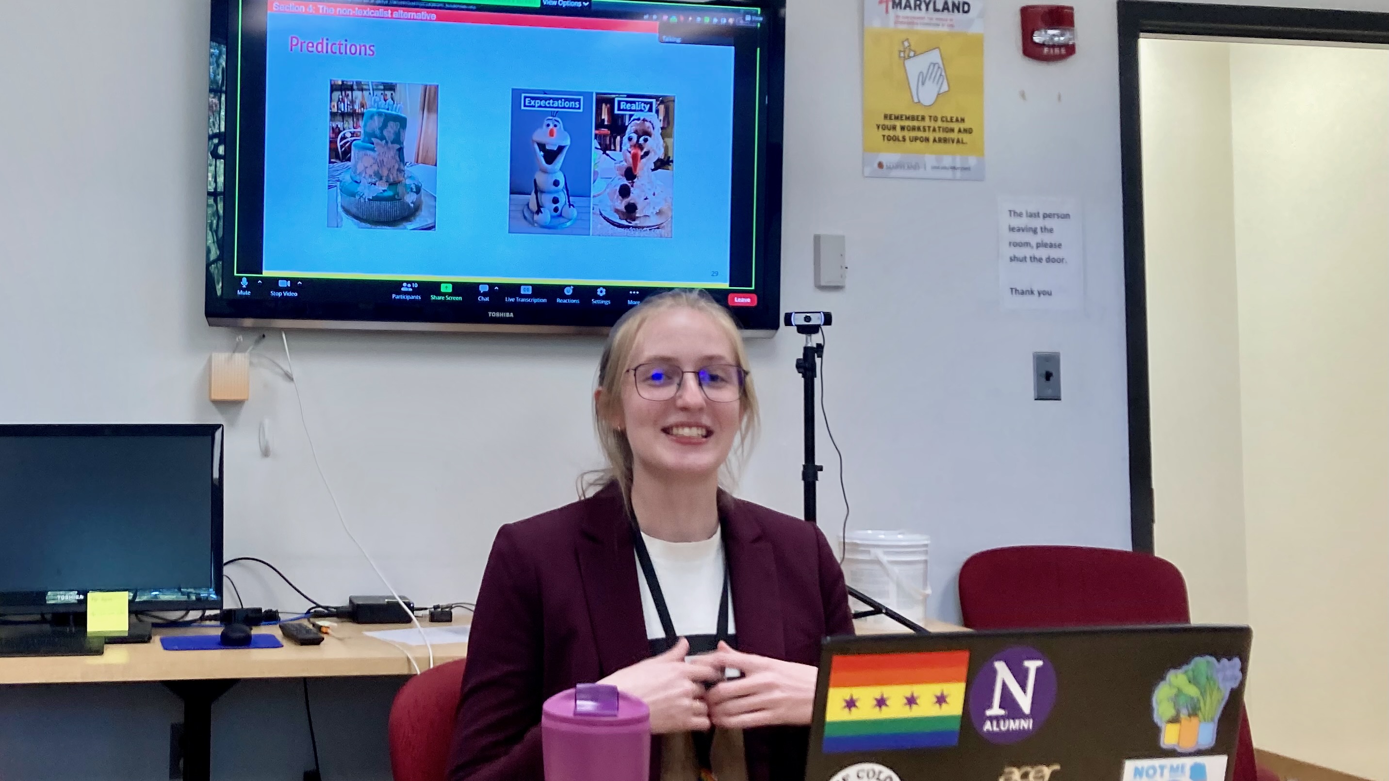 A young woman, sitting at a table smiling, presenting her research, with a tv screen behind her showing a power point slide labeled "predictions", with two photos of elaborate cakes, one tidy and the other a mess.