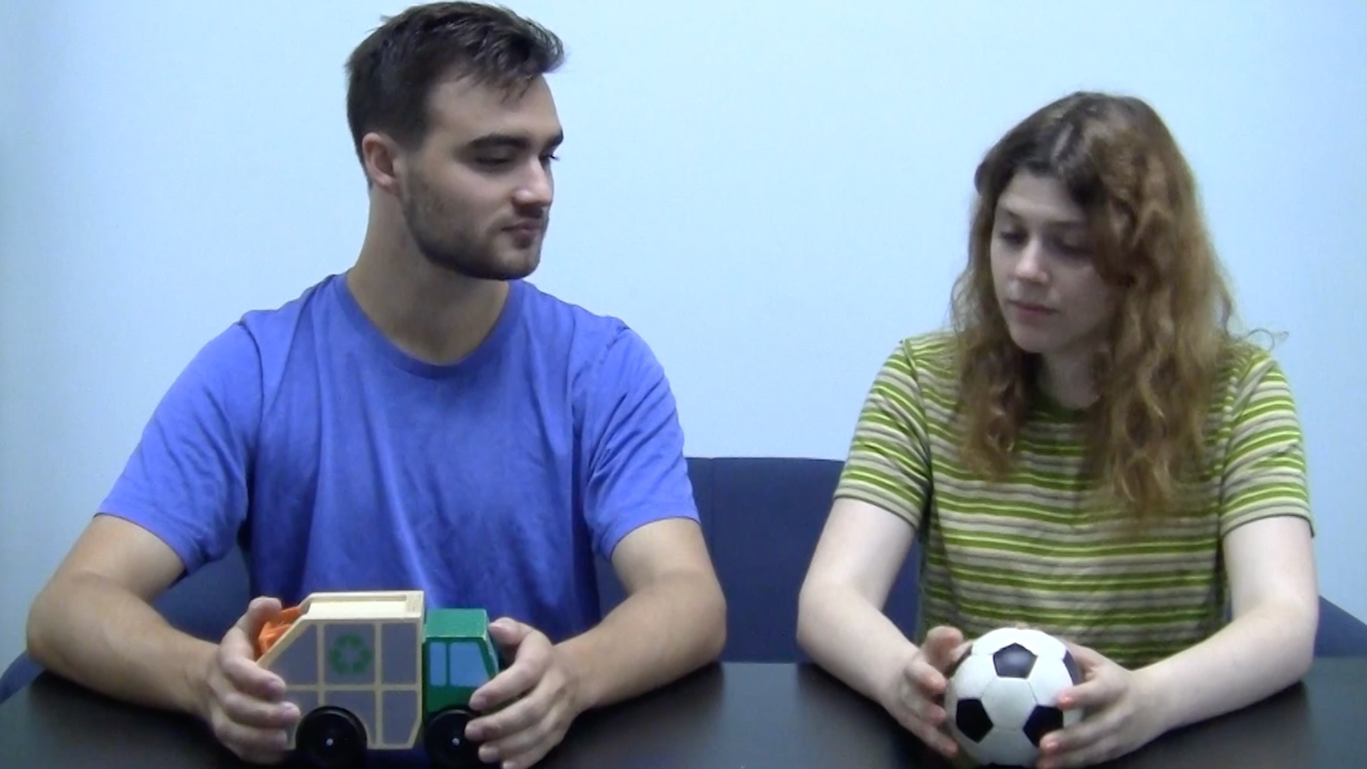 A young man and a young woman sit abreast at a table in an empty room, and calmly exchange a toy garbage truck for a toy soccer ball.