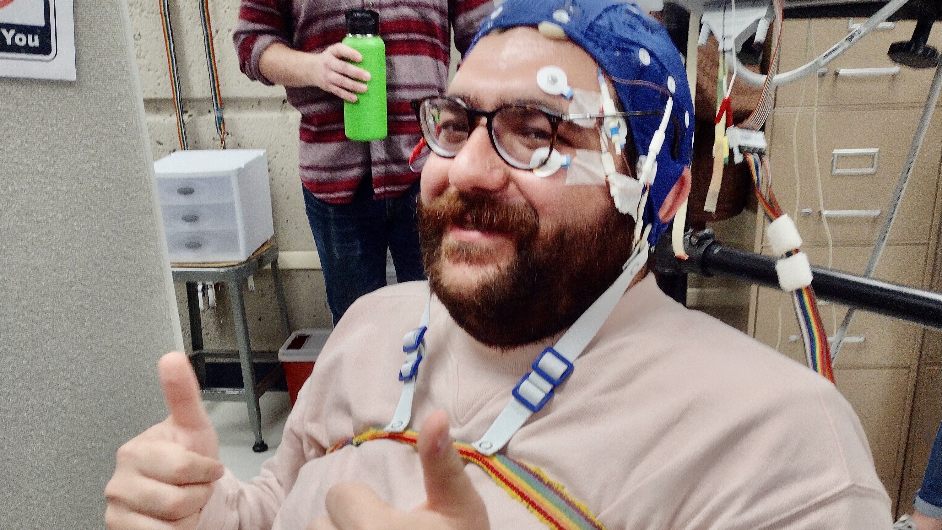 A man with a beard and glasses, with an cap on his head to measure the electrical activity of his brain, giving a double thumbs up.