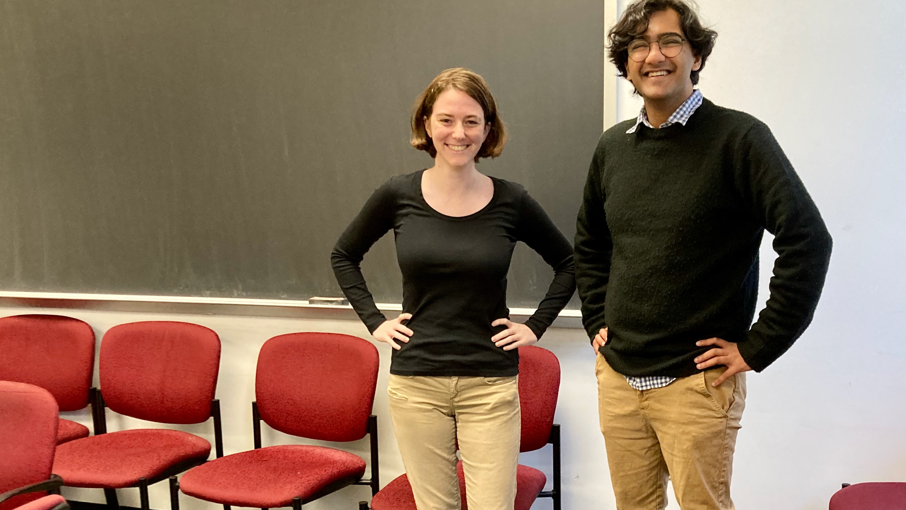 Two classmates, a young woman and a young man, standing side-by-side, arms akimbo, smiling in front of a blackboard, wearing oddly similar outfits: beige pants a black pullover.