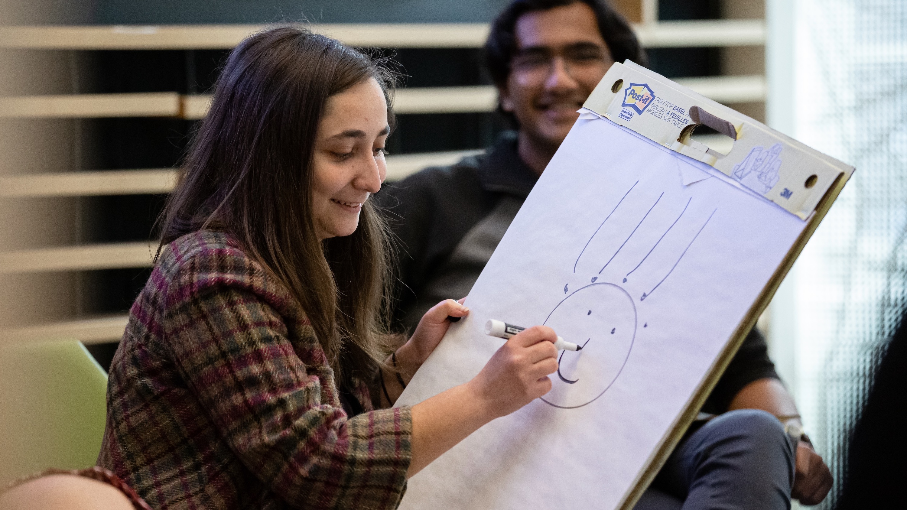 A young woman, Clara Cuonzo, smiling gamely, in a game of pictionary, drawing a person with EEG sensors on their scalp.