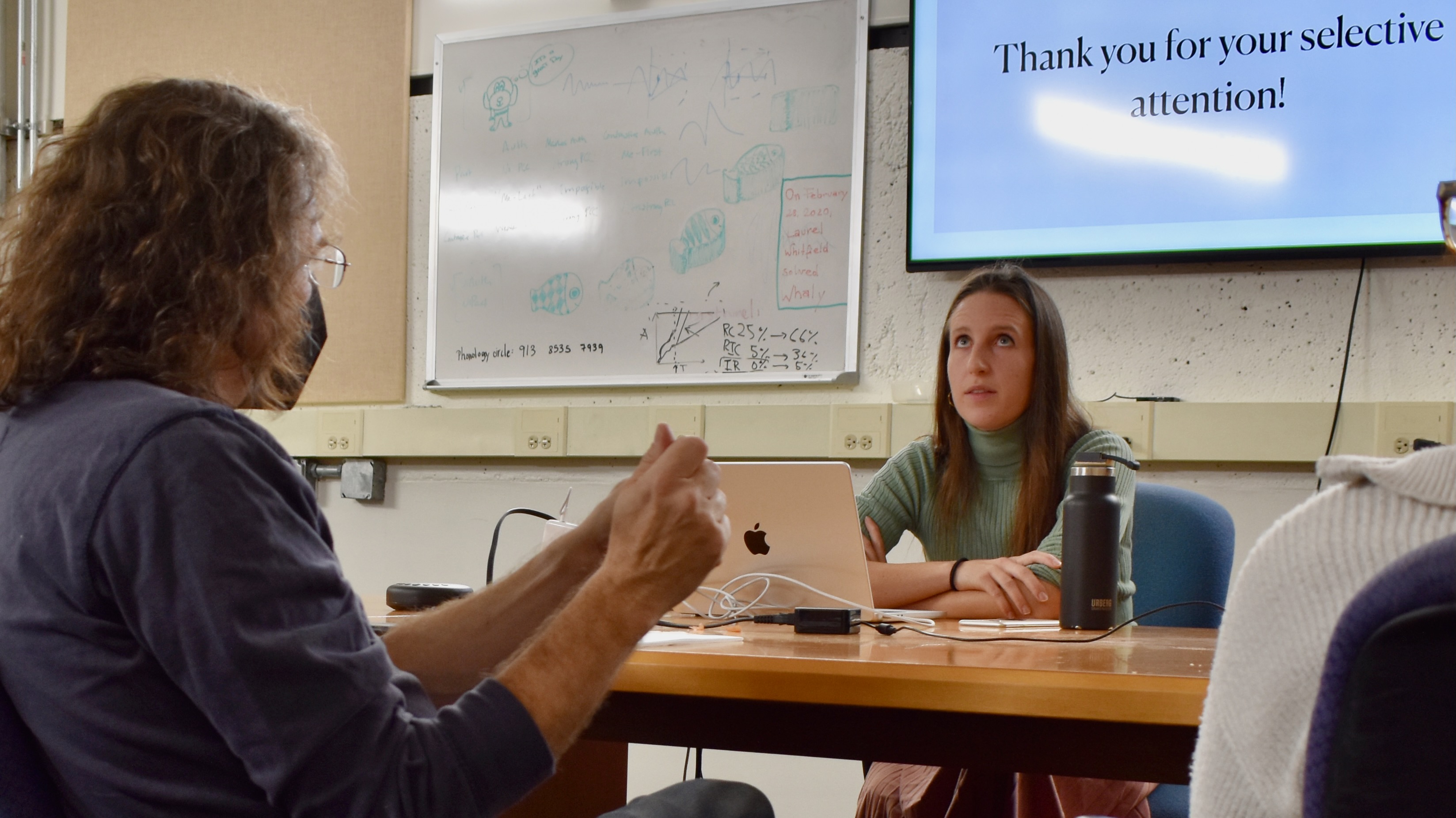 A young woman sitting in front of a tv screen that reads "Thank you for your selective attention", listening to a question from a professor in the foreground.