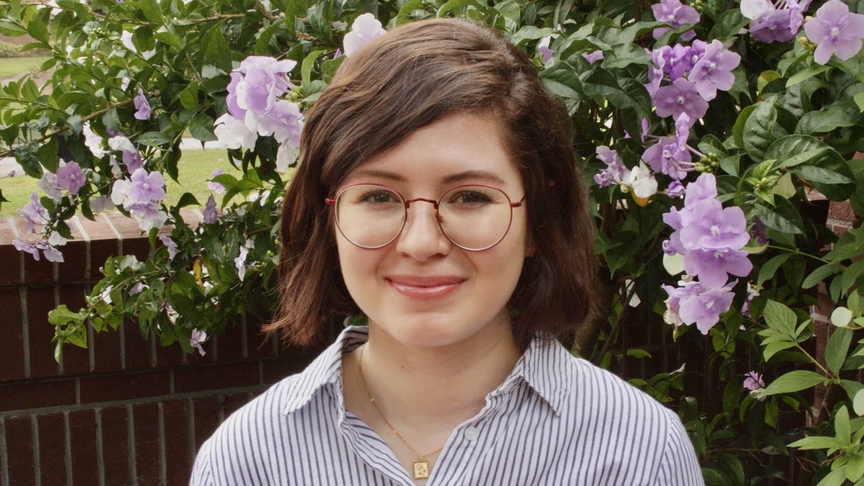 A young woman with a quiet smile, standing outside in the sun, in front of a hedge of lilac flowers.