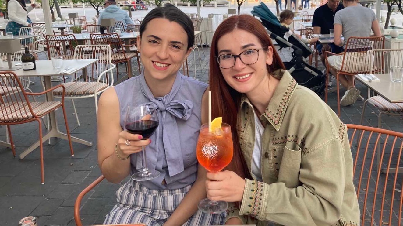 Two young women, seated and smiling at the camera in a sunny piazza, one with brown hair pulled back, the other with auburn hair falling straight down, toasting towards the camera with a glass of red wine and an Aperol Spritz.