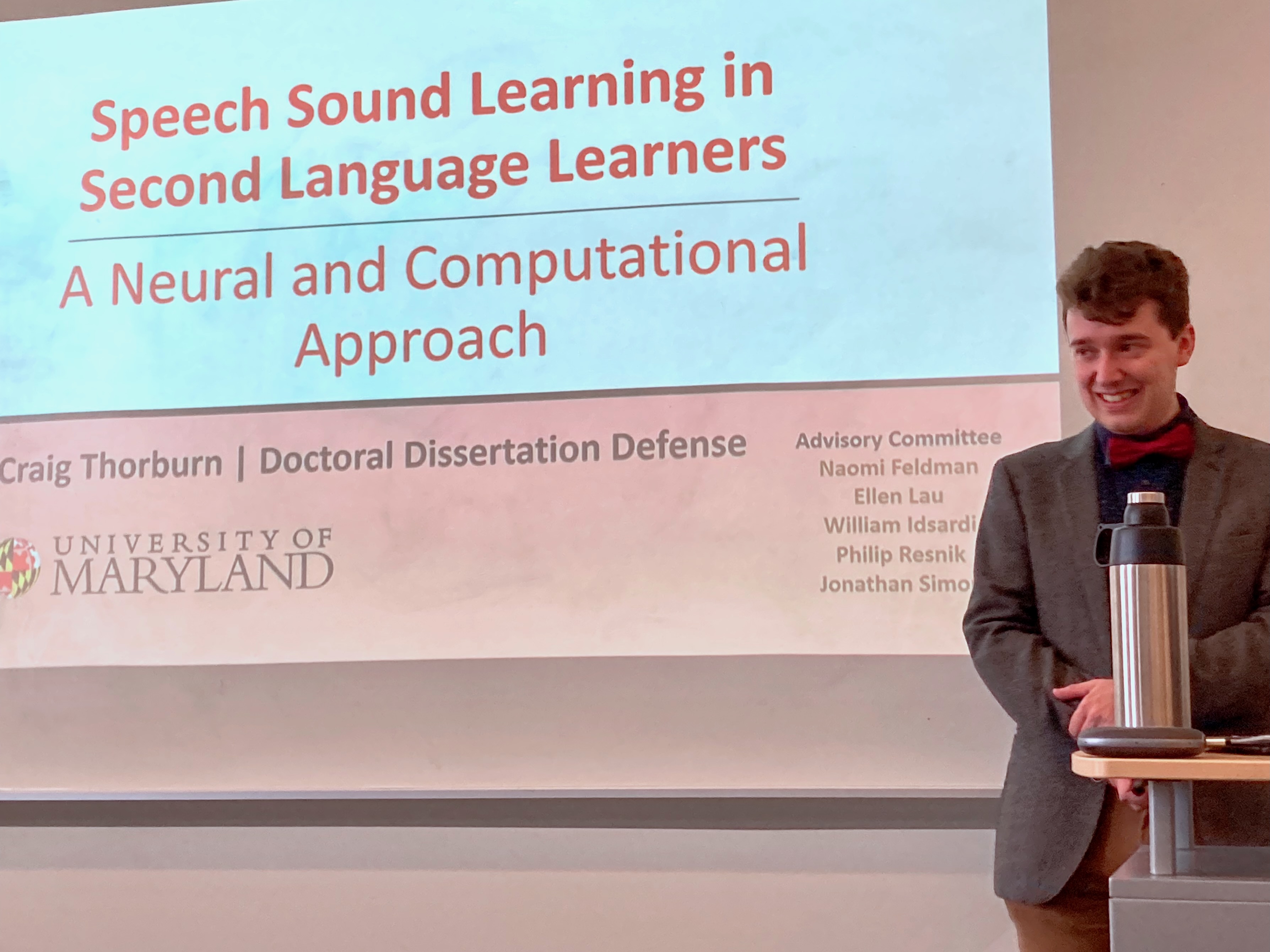 A young man standing nervously in front of a screen, with the title slide of his dissertation defense.